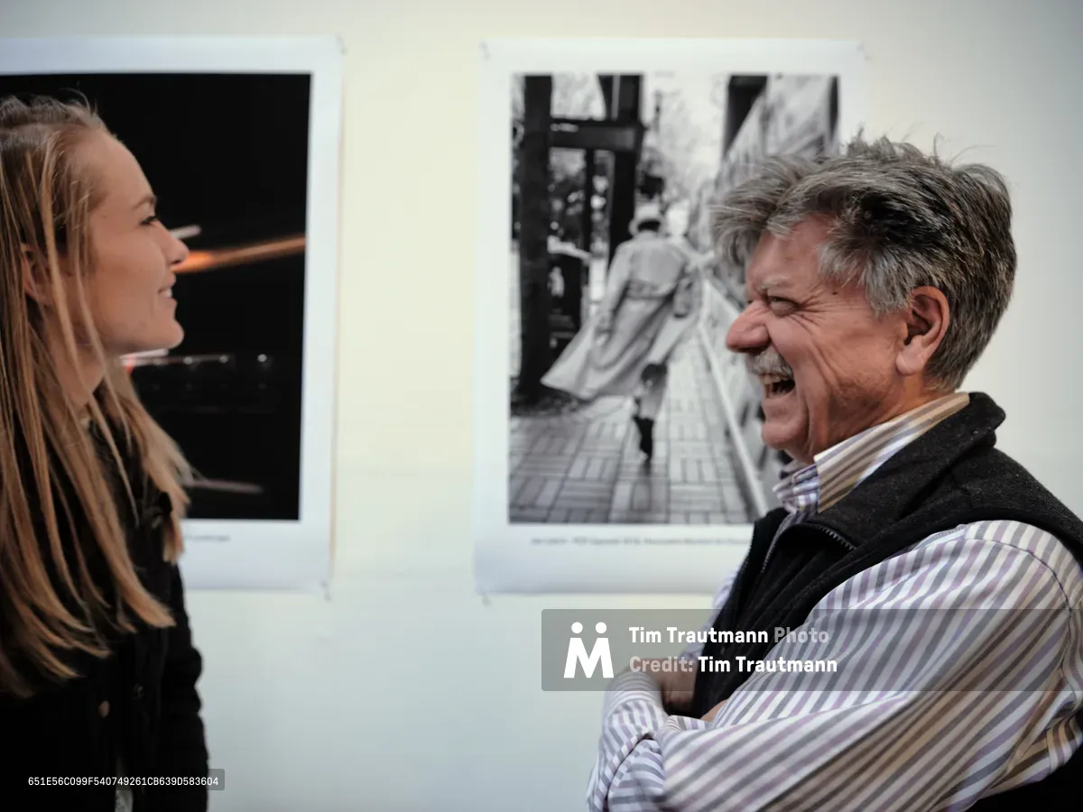 Documentary photographer Joel Leitch erupts in genuine laughter while fellow photographer Alisha Jucevic admires his winning black and white street photograph displayed on the pristine white gallery wall. The intimate moment captures the joy of artistic recognition at ASMP Oregon's PDX Squared 2019 competition, held in Portland's Pearl District. Warm gallery lighting illuminates both the celebrated work and the authentic connection between these creative professionals.