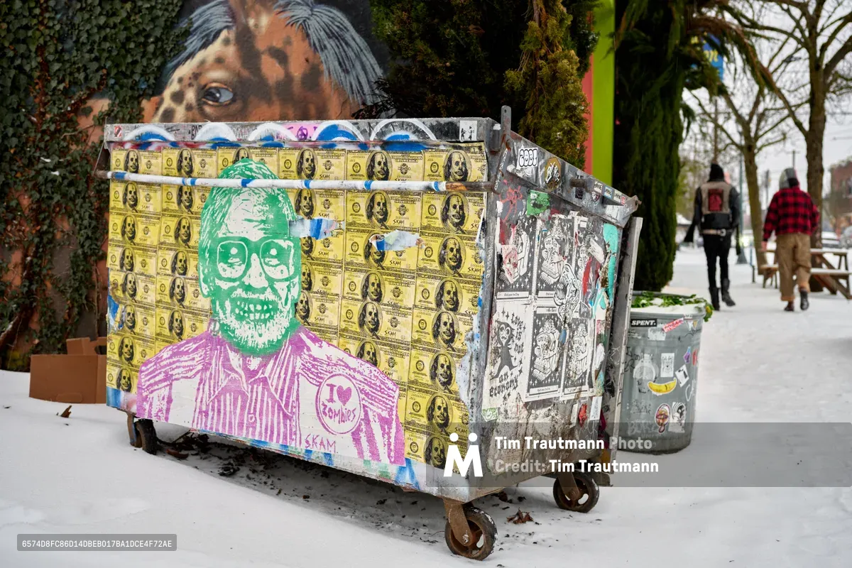 A weathered dumpster on Northeast Alberta Street serves as an anarchic canvas for layered street art, dominated by a striking composition of fake hundred-dollar bills and a green skull wearing glasses above a purple-sketched torso. The winter scene captures Portland's Concordia neighborhood in its raw urban poetry, where the mundane infrastructure transforms into underground galleries. Snow dusts the pavement while pedestrians navigate past this collision of anti-capitalist imagery and guerrilla creativity.