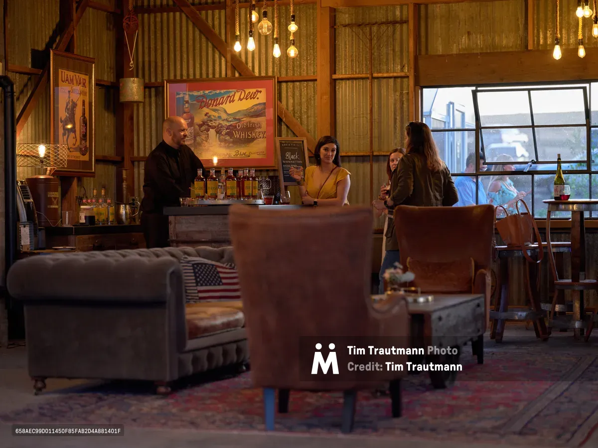 A warmly lit distillery tasting room at Abbey Road Farm in Carlton, Oregon captures an intimate evening gathering. Edison bulb fixtures cast amber light over weathered wood paneling and vintage whiskey advertisements, while a bald bartender in black serves guests at the rustic bar. Visitors examine spirits bottles arranged on the wooden counter as twilight filters through large industrial windows, creating a cozy atmosphere enhanced by leather seating and American flag accents.