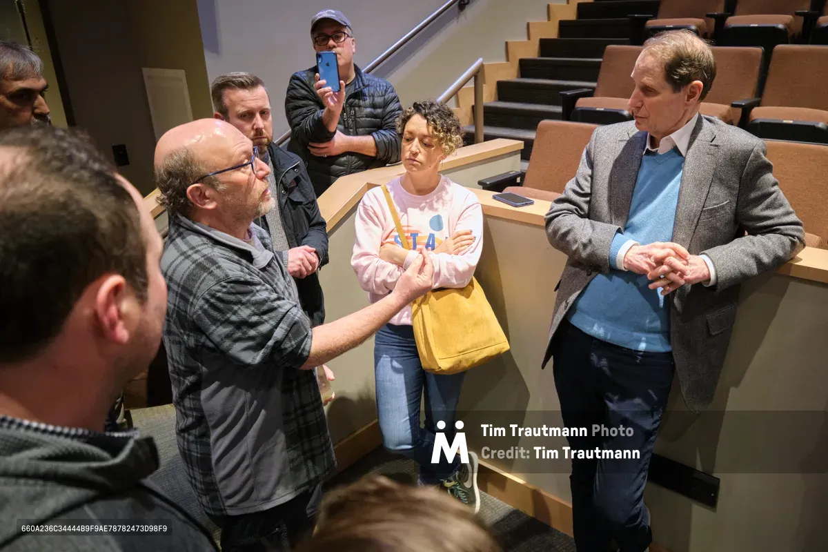 Senator Ron Wyden, distinguished in a gray blazer and blue shirt, leans forward in animated conversation with constituents in a modern lecture hall setting in Portland's Saint Johns neighborhood. The intimate gathering captures the senator's hands-on approach to constituent services, with attendees positioned on tiered wooden seating while one participant records the exchange on their phone. The warm lighting and close quarters create an atmosphere of accessible democracy in action.