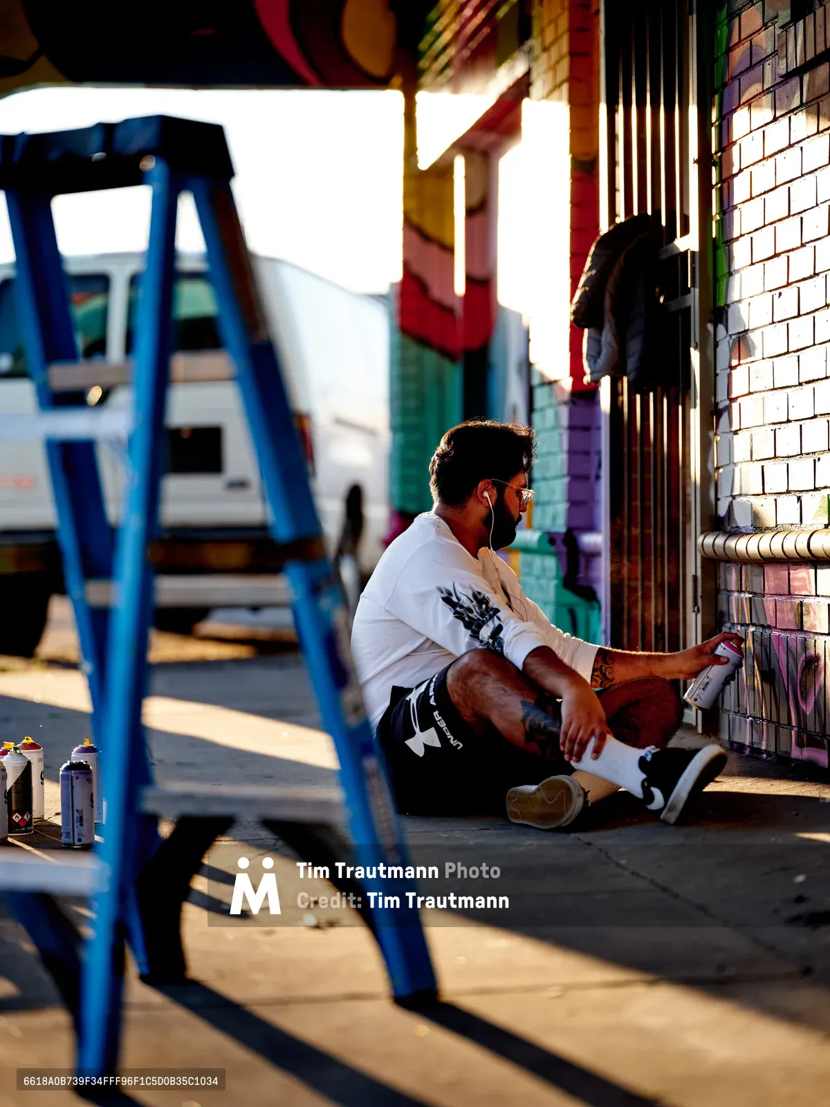 Muralist Heysus crouches in contemplative focus against a vibrant brick wall at the intersection of Northeast 15th and Killingsworth in Portland's Alberta district. Golden afternoon light streams across the scene, illuminating spray paint cans scattered on the concrete while casting dramatic shadows from a blue metal staircase. The artist, wearing a white graphic tee and dark shorts, appears absorbed in his creative process, surrounded by the urban canvas of colorful murals that define this culturally rich neighborhood.