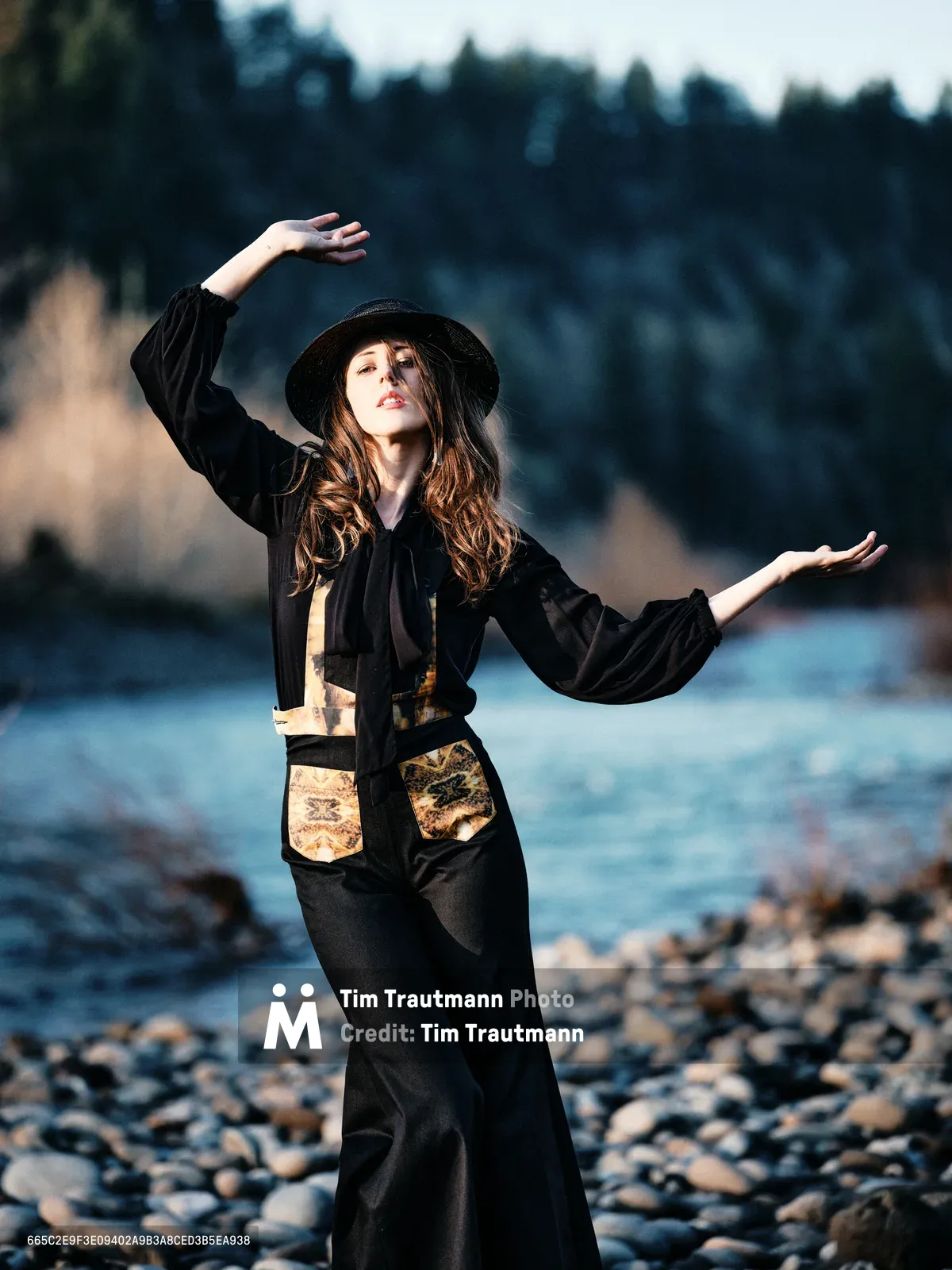 A woman in flowing black attire and wide-brimmed hat strikes an expressive dance pose along the rocky riverbed of Multnomah County's Floodplain Trail. Golden hour light illuminates her auburn hair and ornate vest details while she moves gracefully against the backdrop of the Columbia River and distant evergreen forests. The composition captures a moment of spontaneous movement and connection with Oregon's natural landscape.