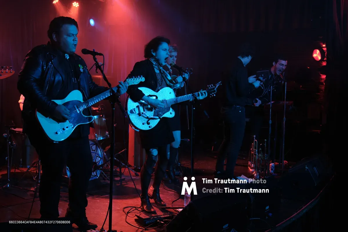 Victor Franco, Charlotte McCaslin, and Faith Grossnicklaus of Roselit Bone perform under saturated red and blue stage lighting at Dante's in Portland, Oregon during RX Fest 2019. The intimate venue's moody atmosphere envelops the trio as Franco commands the microphone on the left, McCaslin centers with her hollow-body guitar, and Grossnicklaus anchors the right side. Theatrical lighting cuts through the smoky darkness, casting the musicians in electric hues while cables snake across the cluttered stage floor.