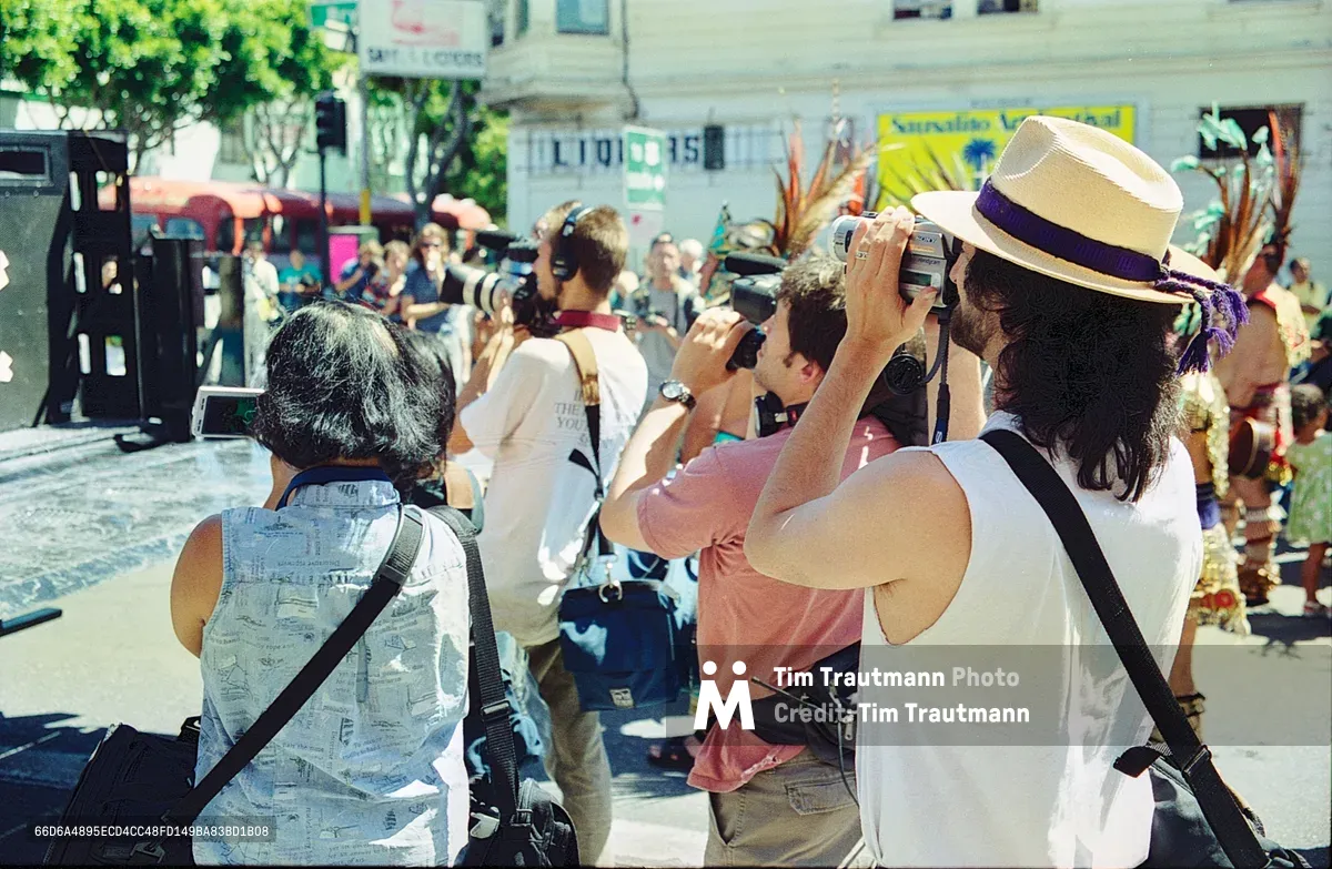 A cluster of photographers and media professionals captures a housing demonstration in San Francisco's Mission District on Bryant Street. The foreground features a woman in a panama hat raising her camera, while colleagues with various recording equipment surround her in the sun-dappled plaza. Behind them, protesters gather with signs and costumes, their voices rising against displacement in this historically Latino neighborhood. The scene pulses with the urgency of gentrification resistance, framed by the district's characteristic low-rise architecture and verdant street trees.