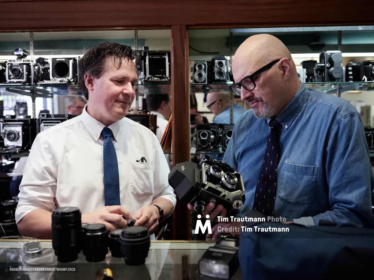 Mike Knight and Arthur Ruckle examine vintage cameras with practiced expertise inside Blue Moon Camera & Machine on North Lombard Street in Portland's Saint Johns neighborhood. Knight, wearing a crisp white shirt and blue tie, handles camera lenses while Ruckle, distinguished in a denim shirt and glasses, inspects a classic camera body. The warm interior lighting illuminates shelves lined with an impressive collection of vintage cameras and equipment, creating an atmosphere of craftsmanship and mechanical precision. Their focused collaboration speaks to decades of shared passion for analog photography and the careful curation that defines this beloved Portland institution.