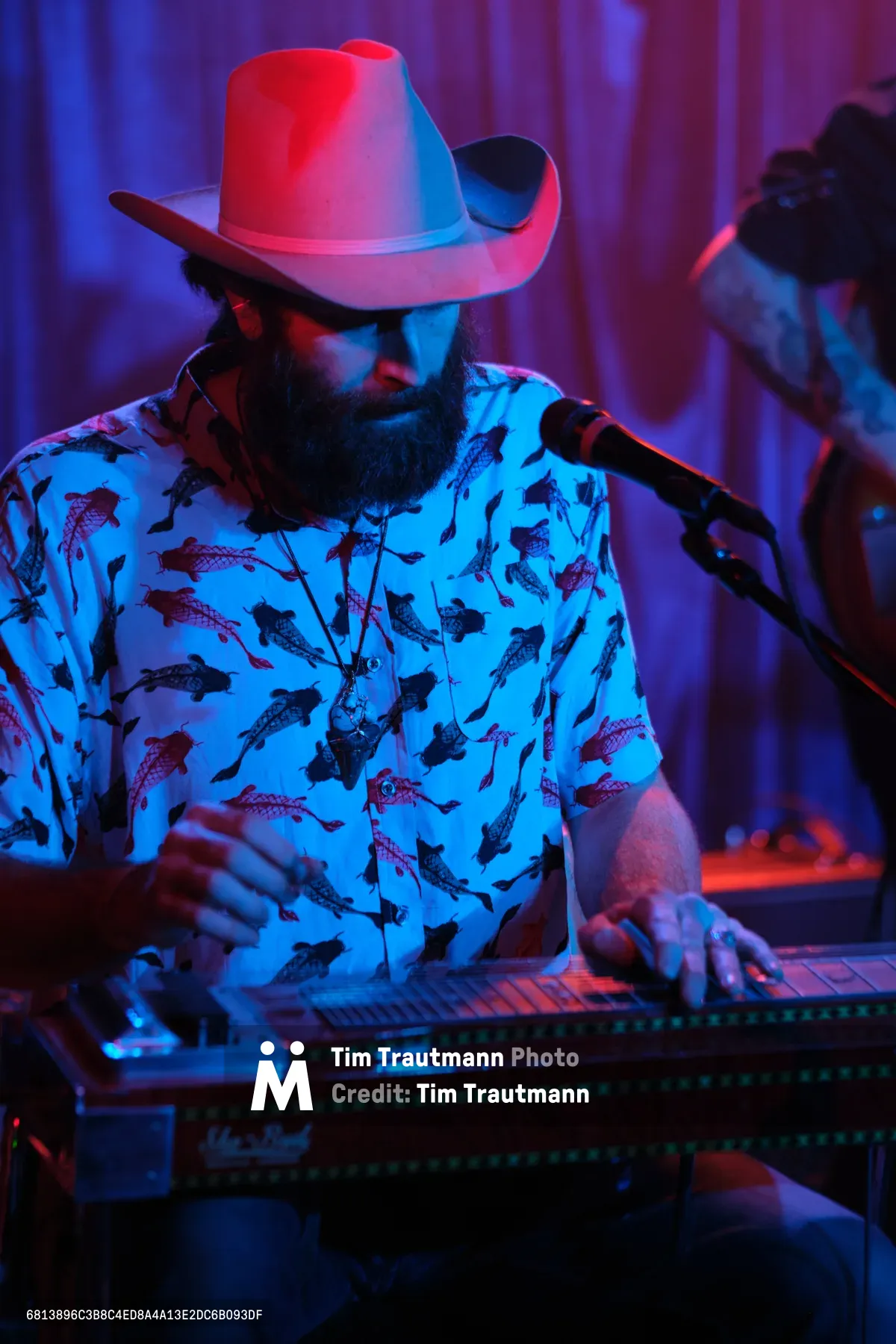 Barry Walker of Roselit Bone commands the intimate stage at Dante's in Portland, his weathered cowboy hat casting shadows over concentrated features as electric blue and magenta stage lights transform the venue into a cyberpunk honky-tonk. His fish-patterned shirt catches the saturated lighting while he leans into his steel guitar, fingers dancing across the strings in a moment of pure musical absorption. The atmospheric lighting creates an otherworldly glow that bridges traditional Americana with futuristic aesthetics, capturing the essence of Portland's eclectic music scene.