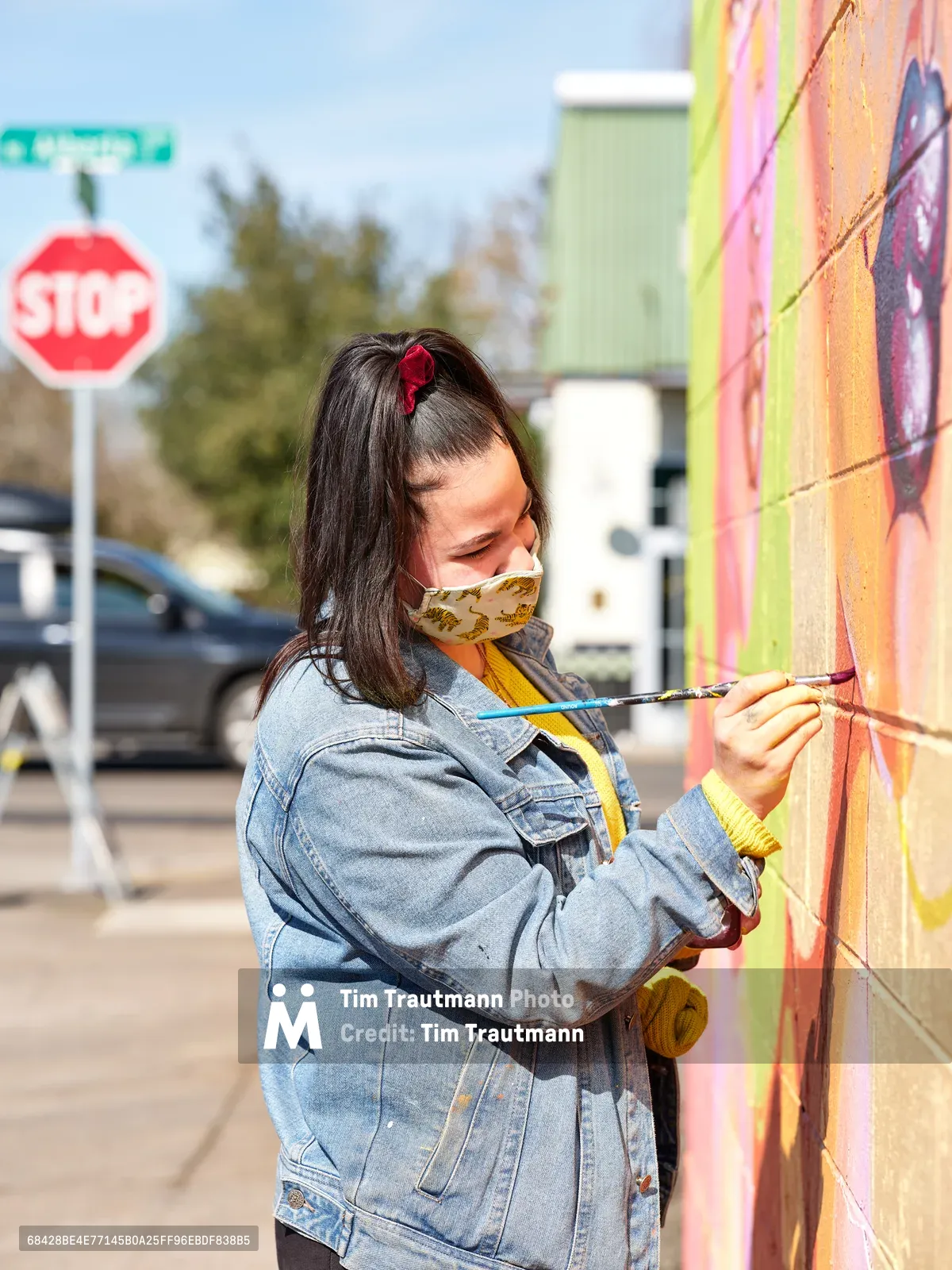 Multidisciplinary artist Vilatose applies delicate brushstrokes to her luminous mural 'Jardín Cubana' depicting three female figures on Northeast Alberta Street in Portland's celebrated arts district. Wearing a paint-splattered denim jacket and protective mask, she works with focused concentration against the vivid coral and lime green wall. The afternoon light catches the wet paint as urban life continues in the background, with stop signs and parked vehicles creating an authentic street art atmosphere.