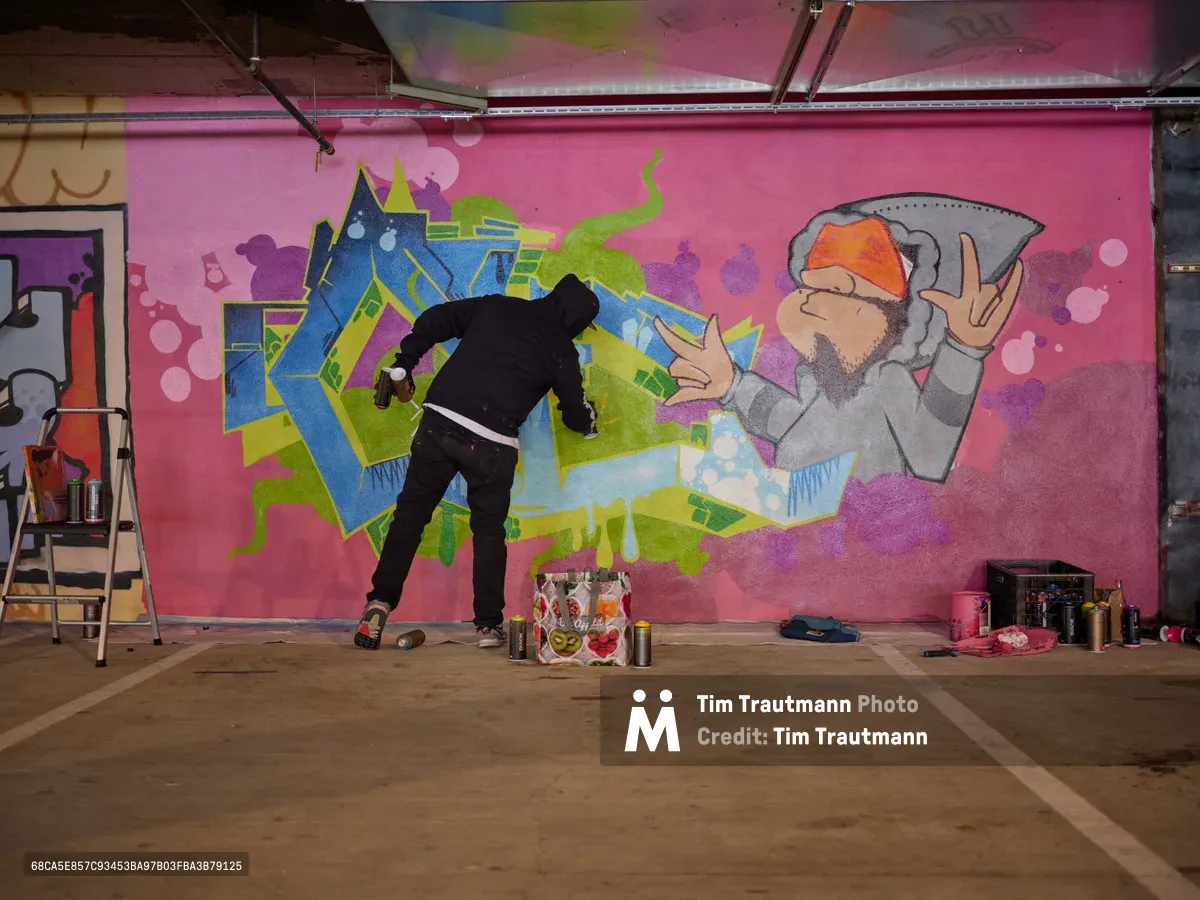 A lone muralist works intently on a vibrant wall painting in the industrial setting of Clay Creative's parking garage in Southeast Portland. The artist, dressed in black, adds details to a fantastical composition featuring geometric shapes, graffiti-style letters, and a whimsical character in a hoodie against a bold magenta background. Spray paint cans and art supplies scatter across the concrete floor, while exposed ceiling infrastructure frames this underground creative sanctuary.