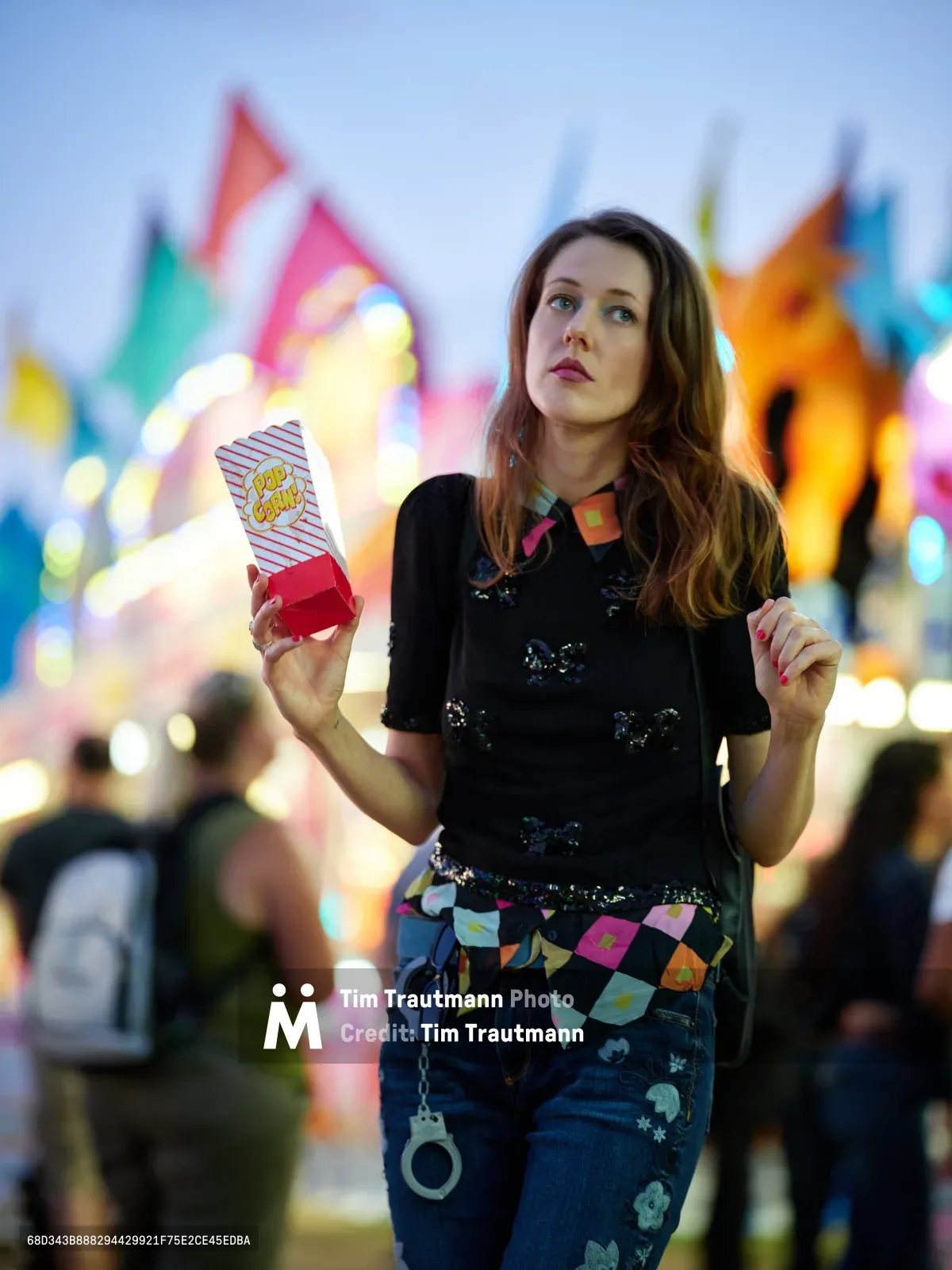 A young woman holds a striped popcorn container against the enchanting backdrop of the Clark County Fair in Vancouver, Washington. Her sequined black top and colorful patterned accessories catch the ambient light while carnival rides blur into a kaleidoscope of warm colors behind her. The evening atmosphere captures the nostalgic magic of county fair festivities, with bokeh lights creating an ethereal glow that frames her contemplative expression.
