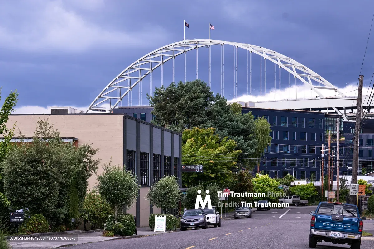 The iconic white steel arch of Portland's Fremont Bridge soars majestically against a dramatic sky of periwinkle and storm clouds, its suspension cables creating rhythmic vertical lines. Viewed from the tree-lined streets of the Slab Town neighborhood, the modernist bridge dominates the urban landscape while leafy deciduous trees and contemporary buildings frame the foreground. Two flags flutter atop the bridge's peak, while parked cars and residential streets below emphasize the human scale against this monumental infrastructure.