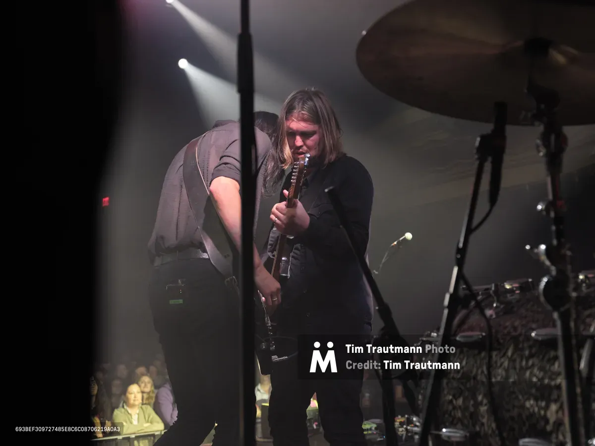 Emma Richardson and Russell Marsden of Band of Skulls perform during their 10 Year Anniversary Tour at Manhattan's historic Webster Hall. Dramatic stage lighting cuts through the smoky atmosphere as Richardson grips her guitar neck while Marsden leans in beside her, both musicians silhouetted against the venue's iconic backdrop. The intimate concert setting captures the raw energy of their decade-spanning musical partnership, with drum kit and amplifiers forming the stage's industrial landscape.