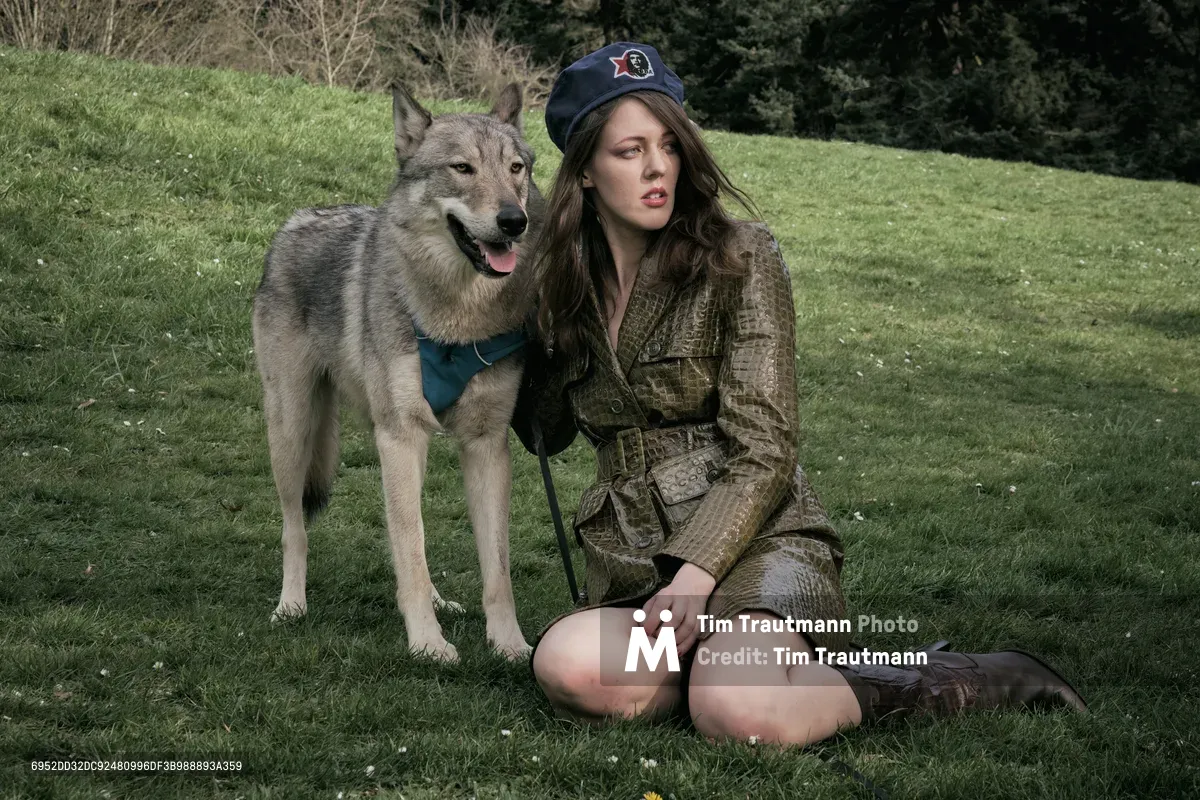 Rhianna Atwood sits cross-legged on the verdant grass of Mount Tabor Park in Portland, accompanied by a majestic wolf-like dog with striking amber eyes and a teal harness. The spring light filters through the park's canopy, creating a moody atmosphere as bare branches frame the background. Her military-inspired olive coat and dark cap create a compelling contrast against the fresh green landscape of this iconic Portland volcanic butte.
