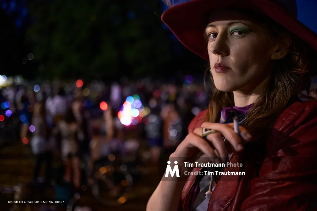 A young woman in a burgundy hat and jacket holds what appears to be a vaping device during Portland's Drop Out Prom Ride, part of the city's annual Pedalpalooza festival. Captured in Colonel Summers Community Garden, the portrait uses dramatic nighttime lighting that illuminates her contemplative expression against a dreamy bokeh of colorful festival lights. The shallow depth of field creates an intimate mood while the blurred crowd and glowing orbs in the background suggest the vibrant energy of the cycling celebration happening around her.