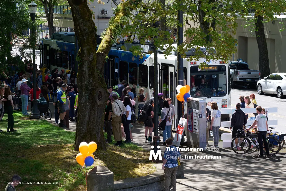 Crowds gather beneath the dappled shade of mature trees along Lloyd Center's sidewalk to witness the retirement of Portland's iconic MAX Type 1 train on a bright April afternoon. Orange and blue balloons punctuate the celebratory scene as transit enthusiasts, families, and community members line the platform area, their shadows creating intricate patterns across the concrete. The white articulated train car displays its familiar blue destination sign while people of all ages document this historic moment in Portland's transportation legacy.