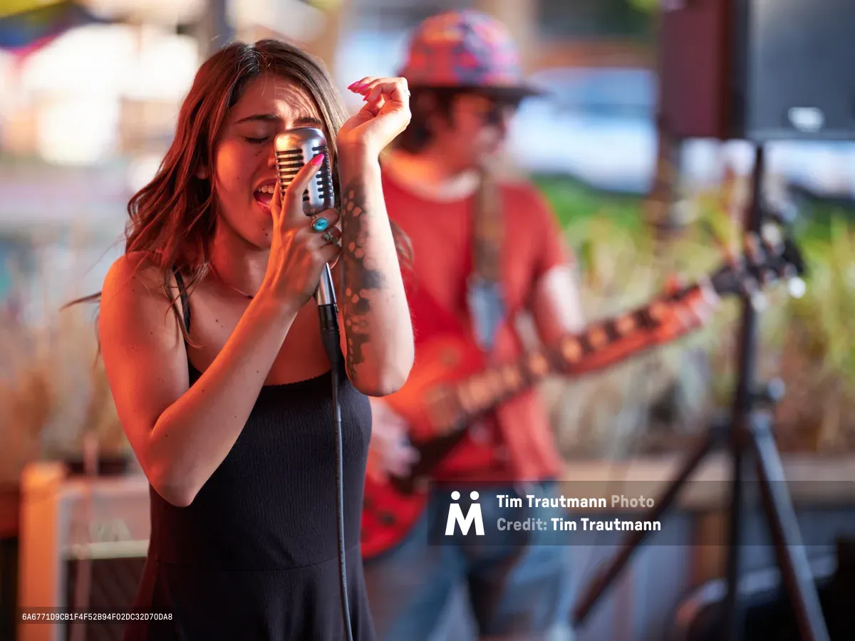 Aubrey Gordon leans into a vintage-style microphone with passionate intensity, her auburn hair catching golden light as she performs at Portland's bustling Mercado. The tattooed vocalist in a sleek black top commands attention while a guitarist in red flannel provides melodic accompaniment in the softly blurred background. Warm evening light bathes the outdoor venue, creating an intimate atmosphere that bridges the energy between performer and audience in Southeast Portland's vibrant cultural hub.
