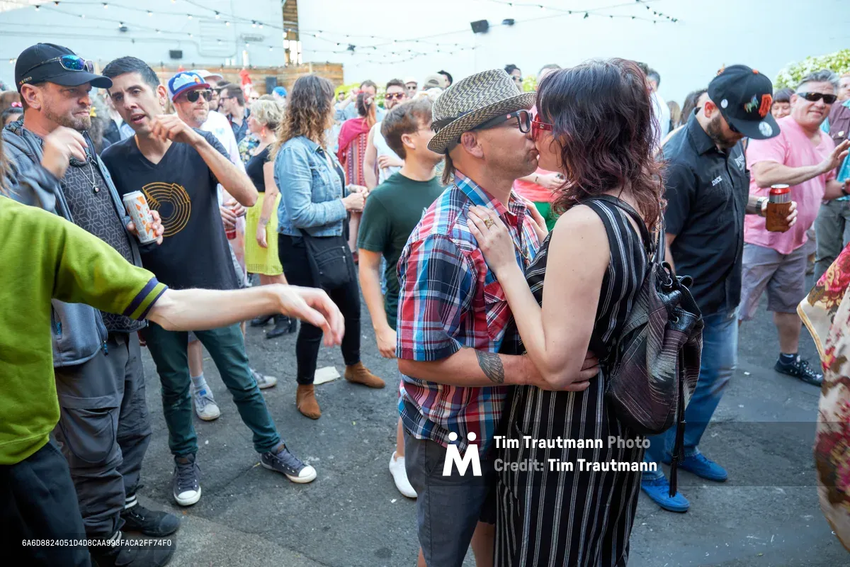 A couple shares a tender kiss in the center of a bustling outdoor day party at White Owl Social Club in Portland. The man in a plaid shirt and tweed cap embraces a woman in pinstripe while dozens of festival-goers mill around them on the asphalt courtyard. String lights overhead and the casual summer attire of attendees create an intimate contrast between private romance and public celebration during this Your Sunday Best season opener.