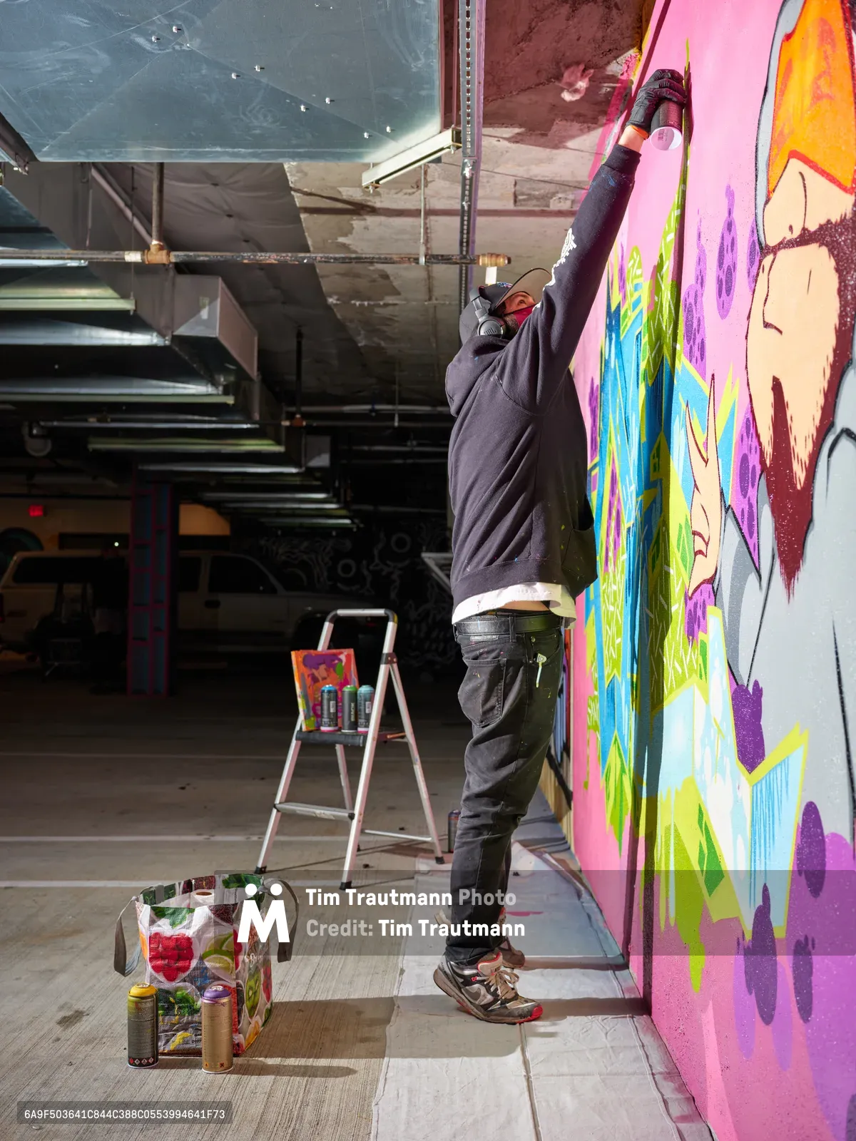 A masked street artist stretches upward to paint a kaleidoscopic mural featuring anime-inspired characters in brilliant pinks, purples, and greens on the concrete wall of Clay Creative's parking garage in Southeast Portland. The industrial space, with its exposed ceiling and metal framework, provides a raw canvas for the emerging artwork. Spray paint cans and art supplies scattered on the concrete floor tell the story of creation in progress, while natural light filters through the urban space.