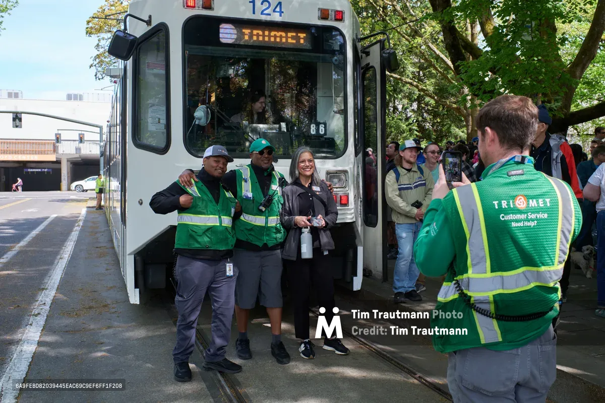 TriMet employees in vibrant green safety vests gather beside the white Type 1 MAX train numbered 124 during its retirement ceremony in Portland's Lloyd District. The scene captures a bittersweet moment as transit workers pose for commemorative photos, their camaraderie evident against the backdrop of leafy street trees and urban infrastructure. Dappled sunlight filters through the canopy, creating a nostalgic atmosphere as onlookers witness the end of an era for Portland's pioneering light rail system.