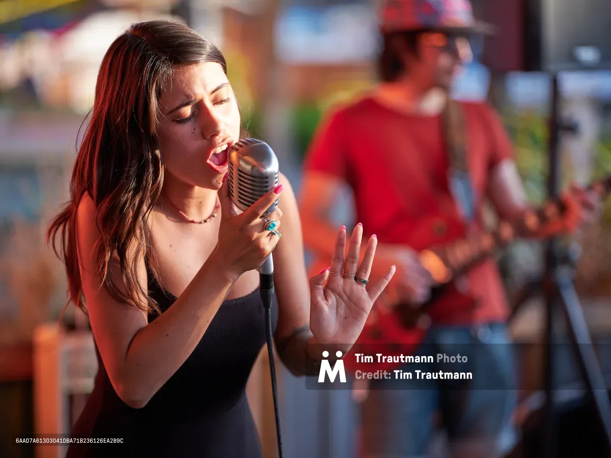 Vocalist Aubrey Gordon delivers an impassioned performance into a vintage-style microphone at Portland Mercado, her eyes closed in musical concentration as golden hour light bathes her expressive face. Her raised hand gestures emphasize the emotional intensity of her song while a blurred musician in red provides atmospheric depth in the background. The Southeast Portland food hall's colorful storefronts create a vibrant bokeh backdrop, capturing the intimate energy of live music in this multicultural community gathering space.