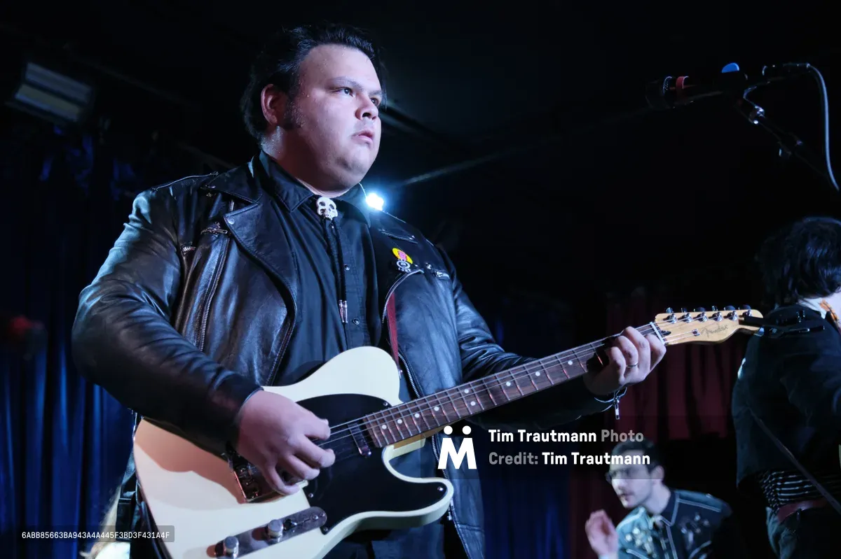 Victor Franco of Roselit Bone commands the dimly lit stage at Dante's in Portland, his cream-colored Telecaster gleaming under moody blue stage lights. Clad in a weathered black leather jacket adorned with pins, Franco's concentrated expression captures the raw energy of underground rock performance. The atmospheric lighting carves dramatic shadows across his face while a bandmate remains partially visible in the background, creating an intimate yet electrifying concert scene.