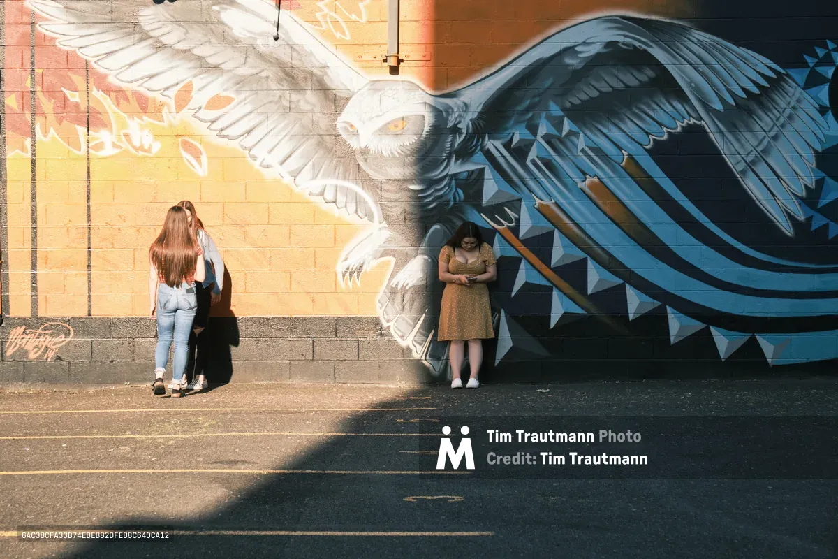 Against the Pearl District's vibrant street art, three figures maintain natural distances along a sun-dappled sidewalk, unknowingly embodying social distancing before the concept entered our collective consciousness. A magnificent owl mural with outstretched wings dominates the brick wall, its piercing gaze overseeing the scene as golden hour light bathes the urban canvas. Two women stand apart examining the artwork while a third figure retreats into shadow, creating an unintentional tableau of personal space and solitary contemplation.