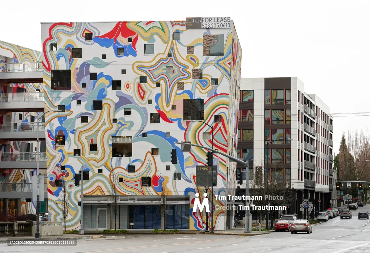 A striking multi-story building in Portland's Buckman neighborhood showcases an explosive psychedelic mural that transforms its entire facade into a swirling kaleidoscope of color. The organic, flowing patterns in vibrant blues, reds, yellows, and pastels cascade across the white surface, punctuated by geometric black squares that serve as windows. The overcast Pacific Northwest sky provides a muted backdrop to this urban art installation, while contemporary apartment buildings and wet asphalt streets frame the scene in typical Portland fashion.