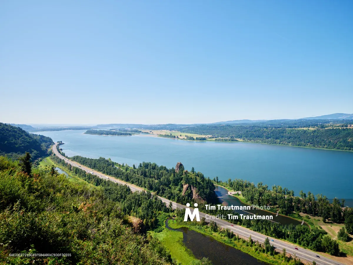 An elevated panoramic view from Vista House captures the majestic Columbia River Gorge as it winds through the Cascade Mountains of Oregon. The serpentine Historic Columbia River Highway traces the southern shoreline below, while emerald forests cascade down volcanic bluffs toward the mirror-still waters. Clear summer light illuminates the pastoral valley stretching eastward, where golden agricultural fields meet distant mountain ridges beneath an azure sky.