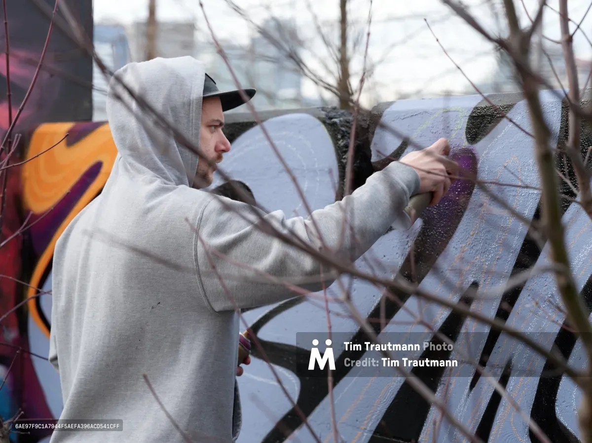 Travis Fields, known as Campographic, carefully applies purple paint to a concrete wall at the Clay Creative Building in southeast Portland's Hosford-Abernethy neighborhood. The hooded street artist works methodically behind bare winter branches that create natural framing across the composition. His gray hoodie contrasts against the textured concrete surface as vibrant orange and purple graffiti elements emerge in the industrial creative space.