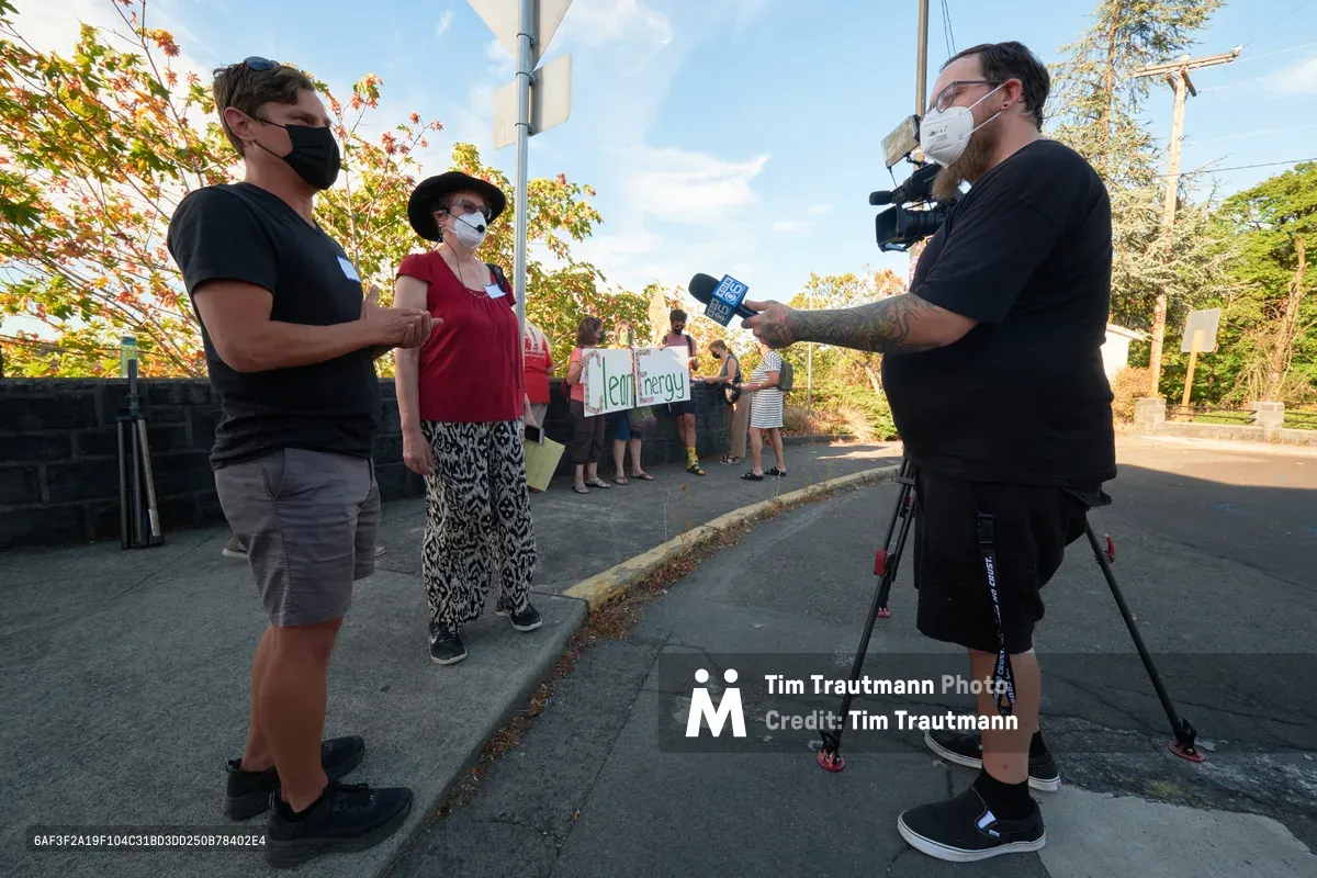 In the golden hour light of McLoughlin Promenade, climate activist Michael Gaskill speaks with a KOIN Channel 6 news crew during a demonstration outside Representative Kurt Schrader's Oregon City office. Masked protesters gather in the background holding signs advocating for clean energy, while autumn foliage frames the scene. The low-angle composition captures the intersection of grassroots activism and media coverage in this suburban political landscape.