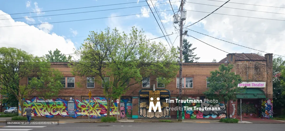 Vibrant murals cascade across the brick facade of the former Oregon Theater building on Southeast Division Street in Portland's Richmond neighborhood. The colorful street art, featuring bold graffiti-style lettering, masked figures, and abstract designs, transforms this historic two-story commercial structure into a living canvas. A lone pedestrian crosses the quiet street under overcast spring skies, while power lines create geometric patterns against the cloudy backdrop. The murals, sponsored by the Portland Street Art Alliance, breathe new life into this cultural landmark along one of the city's most dynamic corridors.