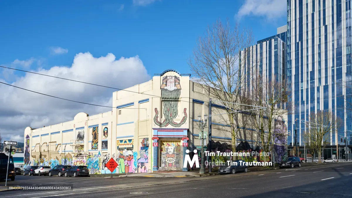 A weathered Art Deco warehouse facade in Portland's Central Eastside district serves as an expansive canvas for vibrant street murals, commissioned by the Portland Street Art Alliance. The cream-colored building displays an eclectic mix of colorful graffiti art across its entire street-facing wall, from abstract designs to character portraits, creating a striking contrast against the gleaming glass towers of the modern cityscape rising behind. Bare winter trees frame the scene under a dramatic sky of billowing white clouds, while parked cars line the quiet urban street.