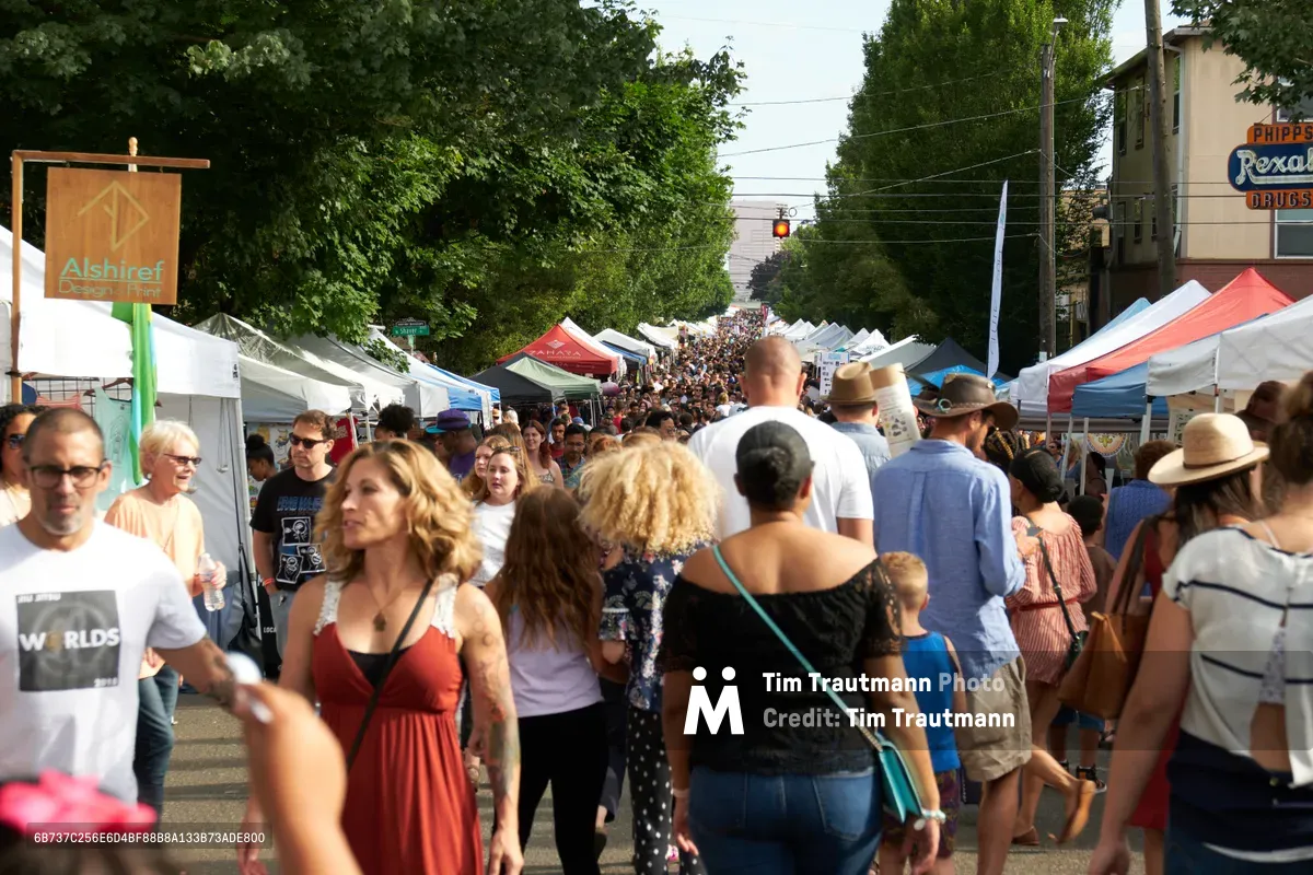 Dense crowds of festival-goers navigate between white vendor tents lining Mississippi Avenue in Portland's Boise neighborhood during a bustling summer street fair. The scene captures the vibrant energy of community gathering, with people of all ages wearing sun hats and casual attire moving through the festival corridor. Lush green trees create a natural canopy over the event, while the warm afternoon light illuminates the diverse crowd exploring local vendors and artisans.