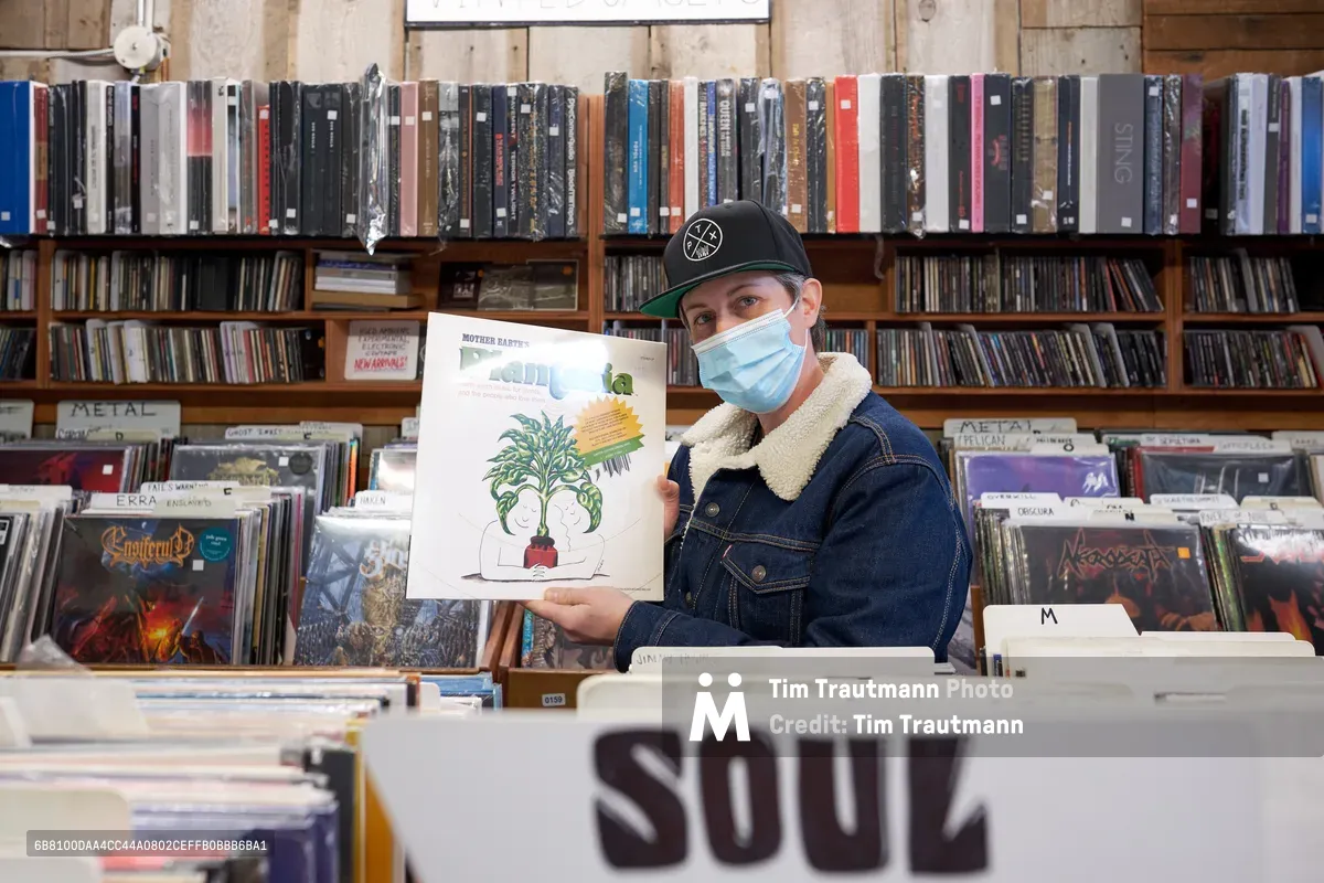 A masked record store employee in a sherpa-lined denim jacket and black cap proudly displays a vinyl album featuring botanical artwork against the towering backdrop of Music Millennium's legendary collection during Record Store Day 2022. The warm, diffused lighting illuminates countless albums organized across wooden shelves that stretch from floor to ceiling, creating an intimate portrait of vinyl culture in Portland's iconic Kerns neighborhood. The scene captures the enduring passion for physical music media during this annual celebration of independent record stores, with alphabetized sections and genre dividers creating a maze of musical discovery.