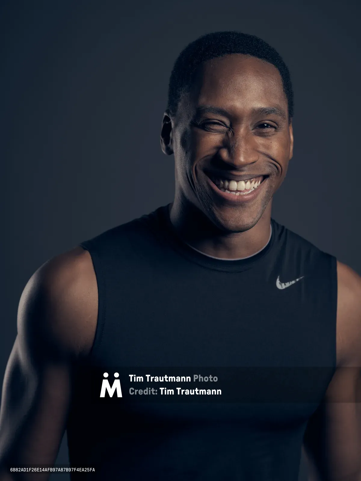 A warmly lit studio portrait captures a Black man in his thirties radiating genuine joy and confidence in Portland's Northwest District. Shot during a medium format camera test session, dramatic side lighting sculpts his features against a deep charcoal backdrop, emphasizing his broad smile and athletic build in a sleeveless performance shirt. The intimate composition draws focus to his expressive eyes and natural charisma, creating a compelling study in contemporary portraiture.