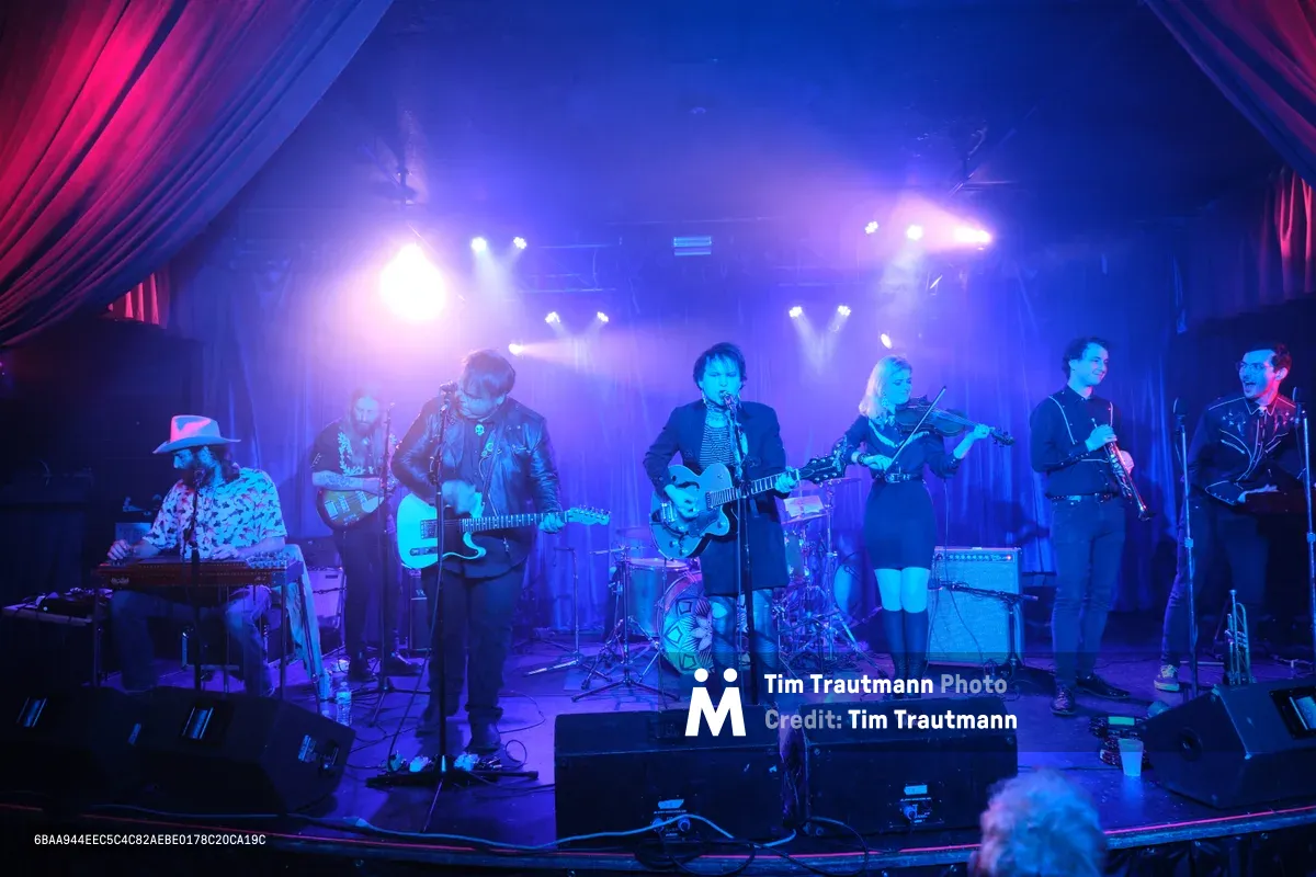 Six musicians command the intimate stage at Dante's in Portland, bathed in electric blue and magenta stage lighting that transforms the venue into a nocturnal dreamscape. The lead vocalist grips a hollow-body guitar while a violinist adds ethereal textures, flanked by keyboards, bass, and drums in this captivating indie ensemble. Crimson curtains frame the performance space as dramatic spotlights slice through the atmospheric haze, creating an otherworldly concert experience.