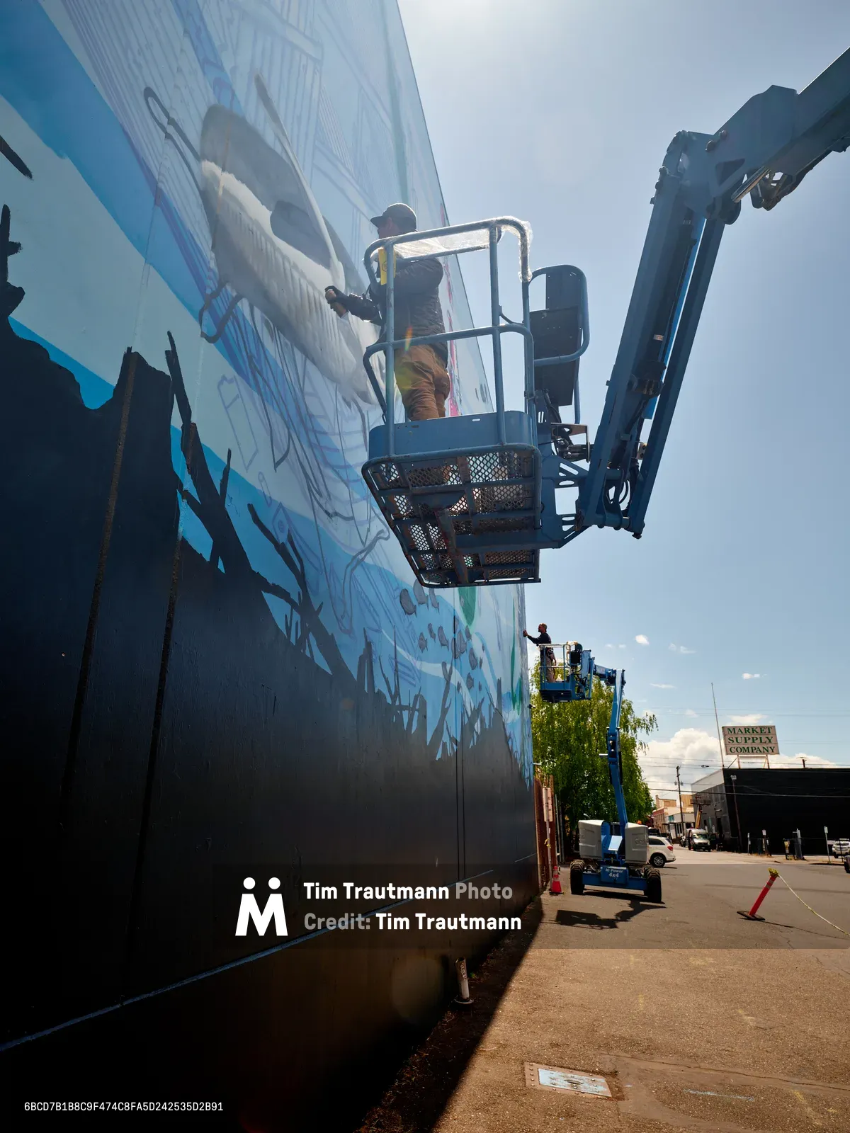Jeremy Nichols and Devin Finley work from the elevated platform of a blue scissor lift, painting a large-scale mural on a building wall in Portland's Central Eastside industrial district. The dramatic low-angle perspective captures the artists suspended against an overcast sky, with vibrant blues and grays emerging across the wall's surface. Industrial buildings and signage frame the background, while safety cones and equipment anchor the urban workspace below.