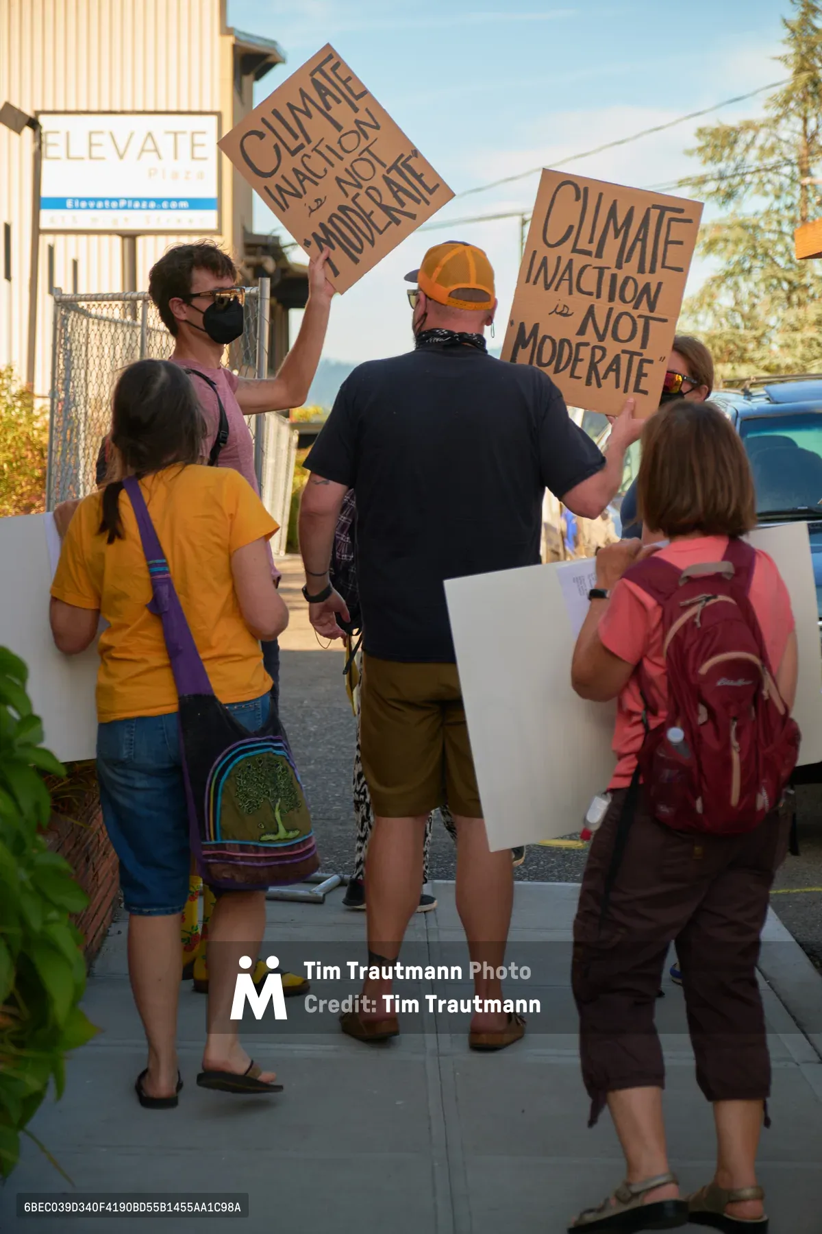 A small group of climate activists gathers on a sun-drenched sidewalk outside Representative Kurt Schrader's office in Oregon City, Oregon, holding handmade cardboard signs demanding climate action over moderation. The golden afternoon light illuminates the diverse group of protesters, including families with children carrying colorful backpacks, as they position themselves near the building's entrance. The scene captures grassroots political engagement in a suburban Pacific Northwest setting, with the activists' casual summer attire and determined postures reflecting the urgency of their environmental message.