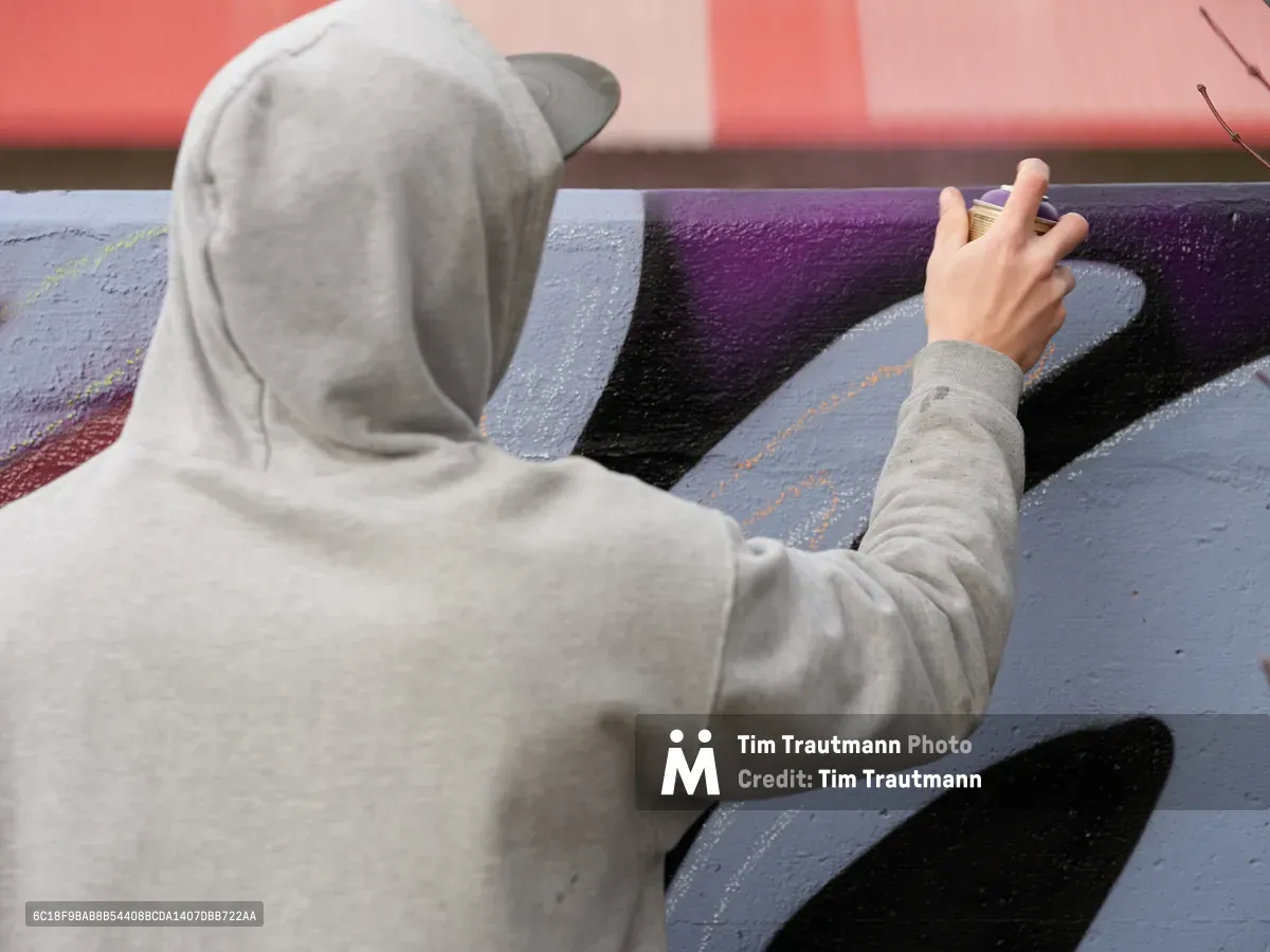 Travis Fields, known as Campographic, works intently on a colorful mural at the Clay Creative Building in Southeast Portland's Hosford-Abernethy neighborhood. Wearing a gray hoodie and cap, the artist applies purple and black paint with focused precision, his outstretched hand guiding the brush across the wall. The warm coral-pink background suggests late afternoon light filtering through the creative space, capturing a moment of artistic dedication in one of Portland's vibrant cultural hubs.