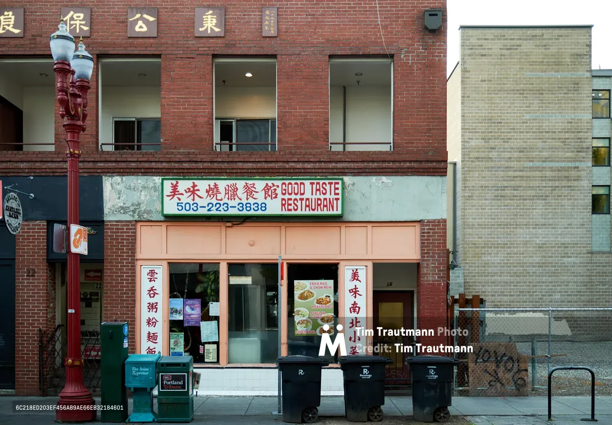 The weathered brick facade of Good Taste Restaurant anchors a corner in Portland's historic Chinatown district, its bilingual signage promising authentic Chinese cuisine beneath golden Chinese characters. Red columns frame the storefront while colorful menu displays peek through windows, creating an inviting gateway to culinary tradition. The urban streetscape reveals the intersection of heritage and commerce, where traditional ornate lamp posts stand sentinel beside modern waste receptacles on the quiet sidewalk.