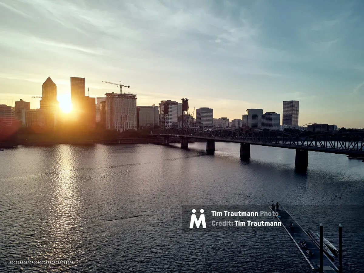 Golden hour light pierces through the Portland skyline as the sun sets behind the downtown towers, casting warm reflections across the Willamette River. The iconic steel truss Hawthorne Bridge spans the water in silhouette, connecting the east and west sides of the city. A small dock extends into the foreground from the Eastbank Esplanade, while construction cranes hint at the city's ongoing growth against the serene evening sky.
