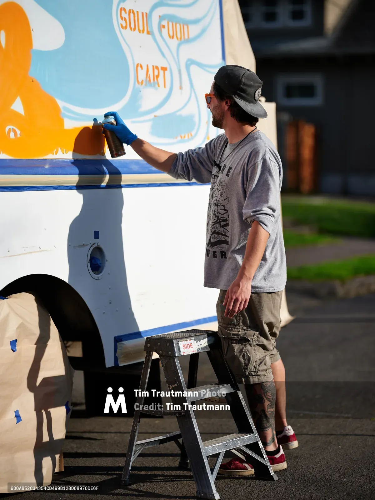 A dedicated muralist in casual work attire and backward cap applies spray paint to the vibrant exterior of a white food truck on a quiet Portland street. The artist works methodically from a metal stepladder, adding finishing touches to colorful graphics that include orange and blue swirls alongside 'SOUL FOOD CART' lettering. Golden afternoon light bathes the scene, casting crisp shadows across the asphalt while residential buildings blur softly in the background.