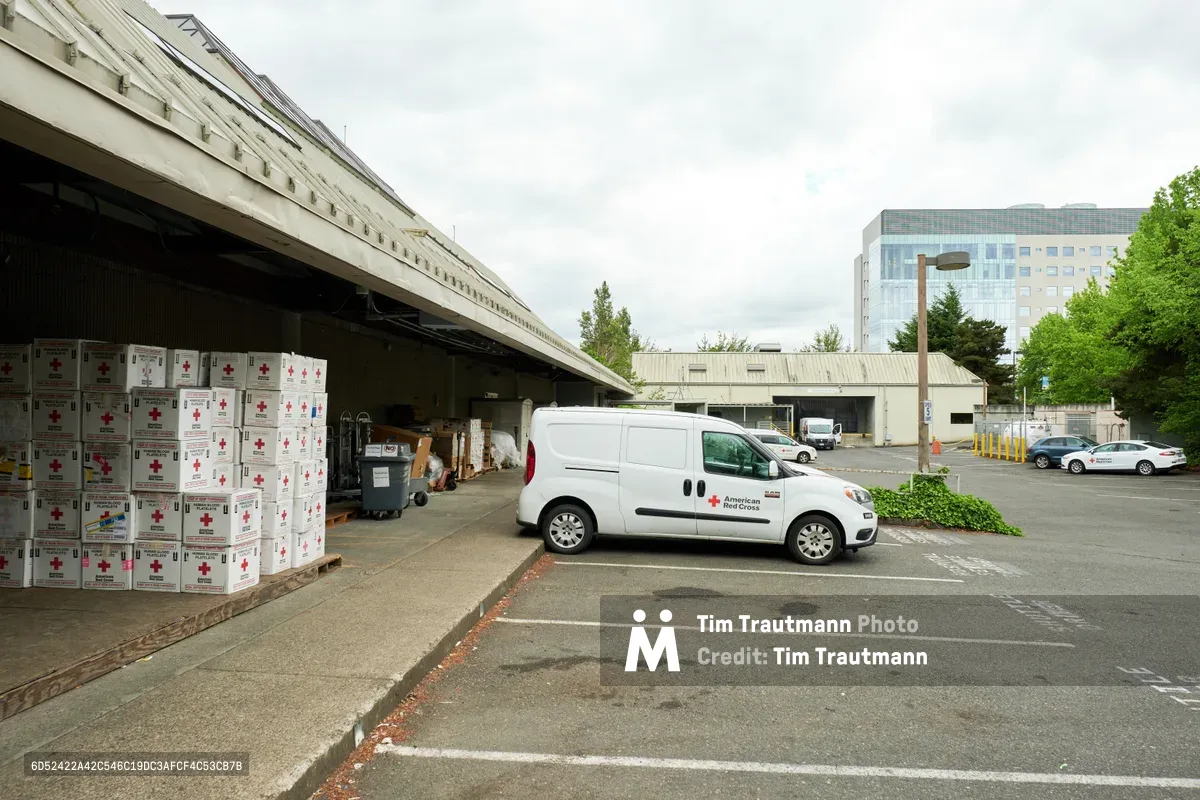 Under overcast Pacific Northwest skies, a white American Red Cross delivery van sits positioned at a concrete loading dock in the organization's Cascades Region facility on North Vancouver Avenue. Towering stacks of empty blood platelet boxes marked with red crosses await collection inside the shadowed warehouse bay, while the modern glass facade of a Portland office building rises in the background. The utilitarian scene captures the operational backbone of humanitarian aid distribution in Oregon's largest city.