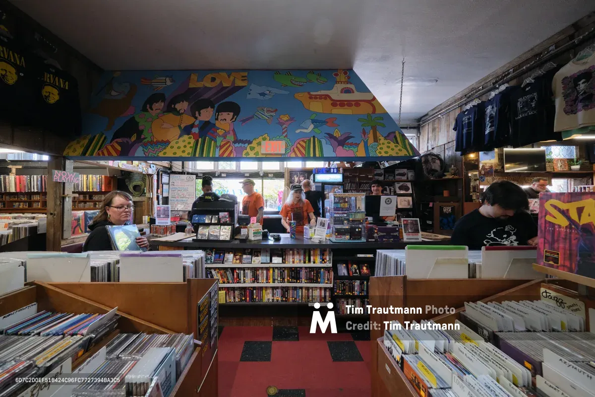 Inside Portland's legendary Music Millennium record store on East Burnside, customers browse through wooden crates filled with vinyl records beneath a vibrant ceiling mural celebrating music culture. The intimate shop buzzes with activity as a woman in glasses examines an album cover while a young man in black searches through adjacent bins. Natural light filters through the storefront windows, illuminating the checkerboard floor and towering shelves packed with albums, CDs, and music memorabilia that define this beloved independent music sanctuary.