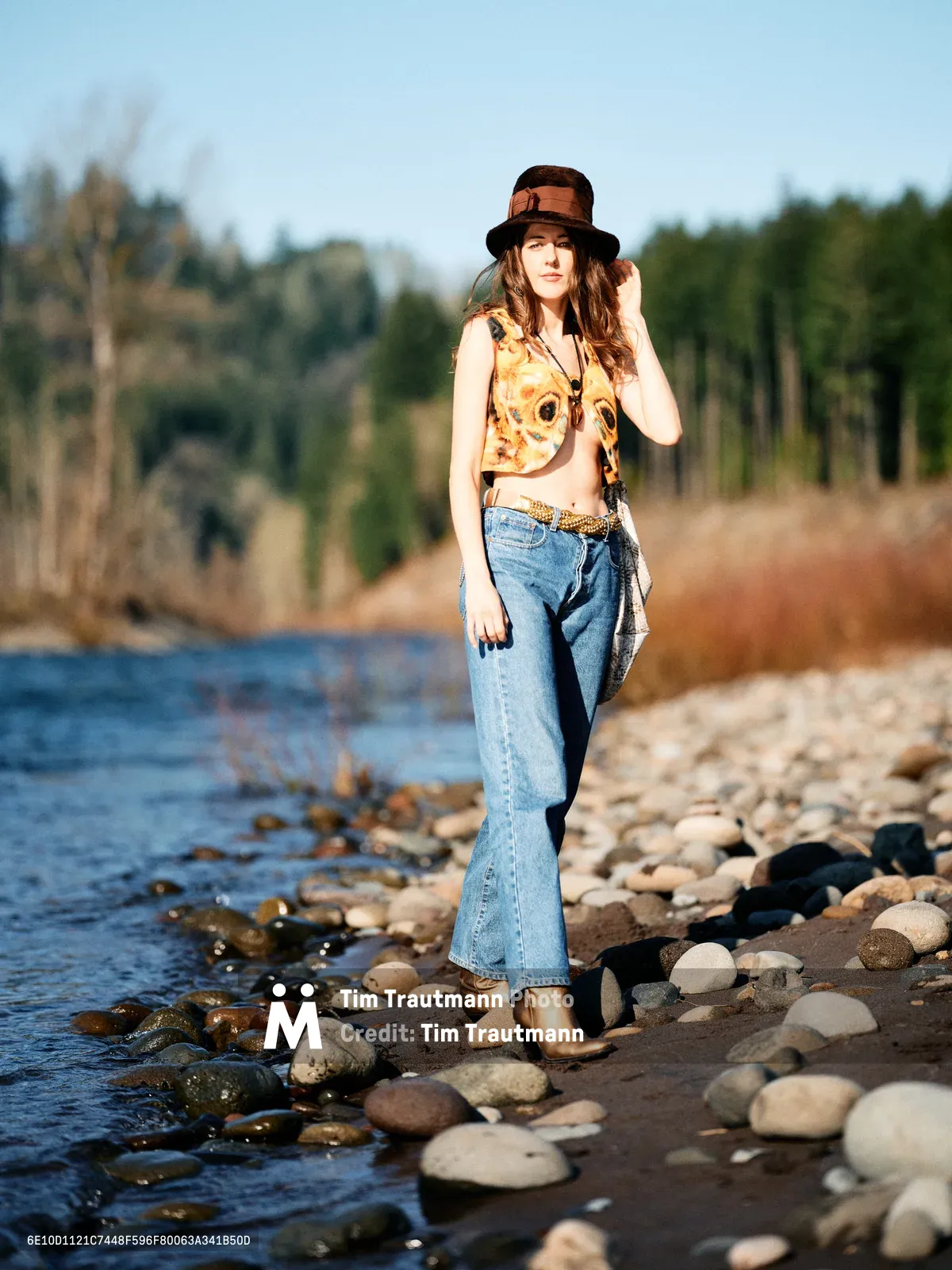 A young woman in vintage-inspired attire poses confidently along the rocky shoreline of the Columbia River near the Floodplain Trail in Multnomah County. Bathed in warm golden hour light, she wears a floral crop top, high-waisted jeans, and a dark fedora while standing among water-smoothed stones. The soft focus background reveals the river's gentle current and dense Pacific Northwest forest, creating an intimate portrait that captures both personal style and Oregon's natural beauty.