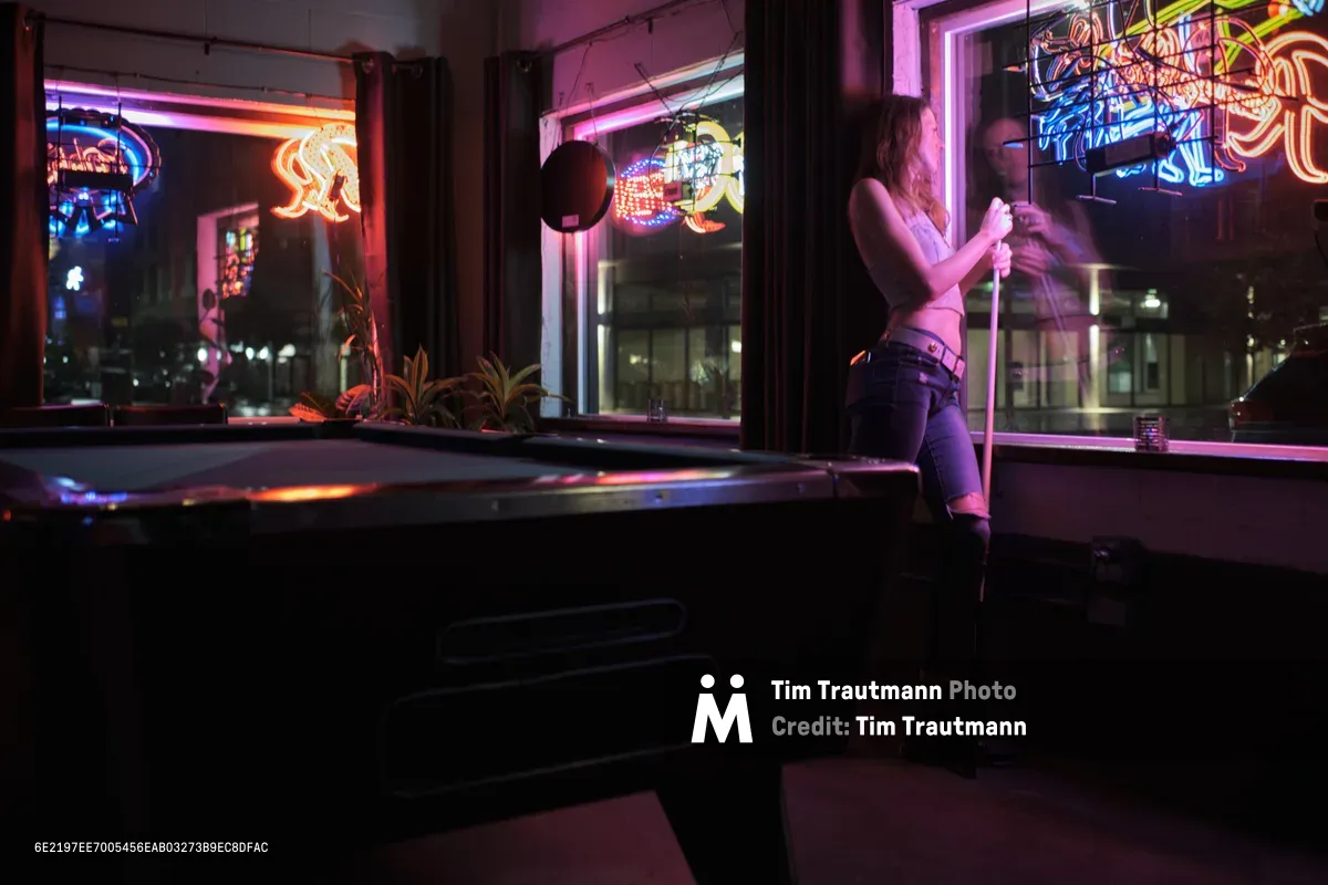 A solitary woman leans against a pool table in the atmospheric interior of Vendetta bar on Northeast Skidmore Street in Portland's Humboldt neighborhood. Electric neon signs cast vibrant pink and orange light through the windows, creating dramatic reflections on the polished surface while she holds a pool cue in contemplative silence. The moody lighting transforms the empty venue into a cinematic tableau of urban nightlife isolation, with the glow from multiple neon advertisements painting the scene in saturated hues.