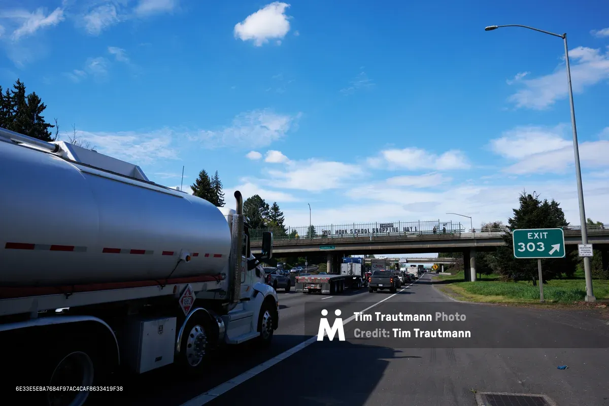 A white tanker truck dominates the foreground as traffic flows beneath a highway overpass on Interstate 5 in North Portland, where activists have displayed a protest banner reading 'No Work School Shopping May 1st.' The scene unfolds under a brilliant blue sky dotted with white clouds, with the green Exit 303 sign marking the location near the Skidmore Street interchange. Evergreen trees frame the composition while the banner stretches across the concrete bridge structure, creating a stark juxtaposition between commercial transportation and grassroots activism.