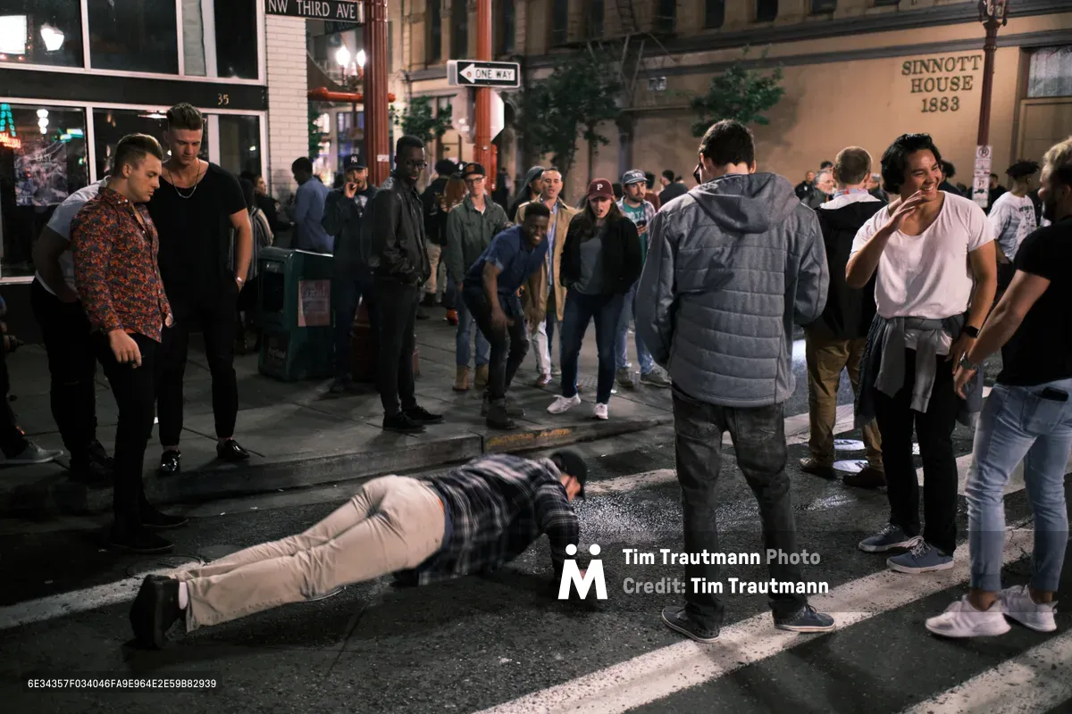A late-night street scene at the corner of NW Third Avenue in Portland's Chinatown as bars and clubs close for the night. A young man in a plaid shirt and khaki pants does push-ups in the middle of the wet street while a crowd of onlookers gathers around him, some laughing and watching with amusement. The historic Sinnott House building from 1883 is visible in the background, along with a one-way street sign and the glow of bar lights on the rain-slicked sidewalk.