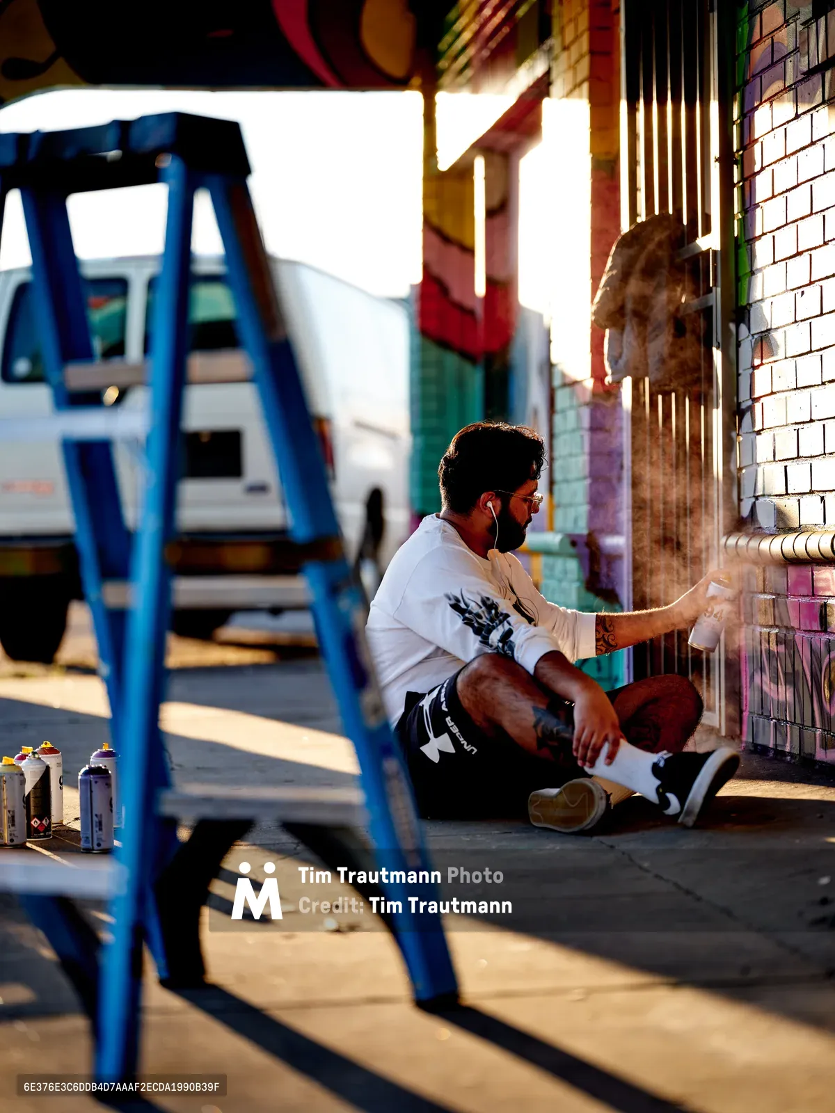 Muralist Heysus sits cross-legged on the sidewalk, painting a vibrant mural on a white-tiled wall in Portland's Alberta neighborhood. Golden hour light bathes the scene, casting long shadows from a blue metal ladder positioned nearby. Spray paint cans scattered on the ground reveal his working process, while the artist focuses intently on adding colorful details to the emerging artwork at the intersection of Killingsworth and Northeast 15th Avenue.