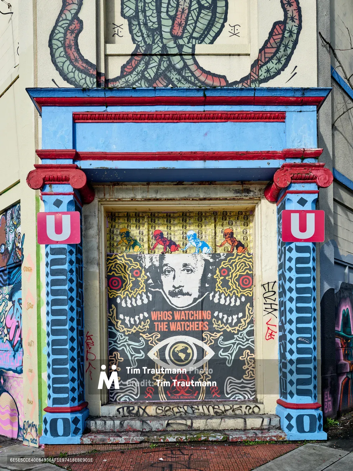 A striking street mural adorns the entrance of a former Salvation Army warehouse in Southeast Portland's Central Eastside district. The artwork features a classical architectural frame painted in vibrant blues and reds, housing a provocative Orwellian-themed piece with Einstein's portrait and the text "Who's Watching the Watchers" beneath an all-seeing eye. Serpentine murals twist above the doorway while additional street art layers the surrounding concrete walls, creating a powerful commentary on surveillance and authority within this transformed industrial space.