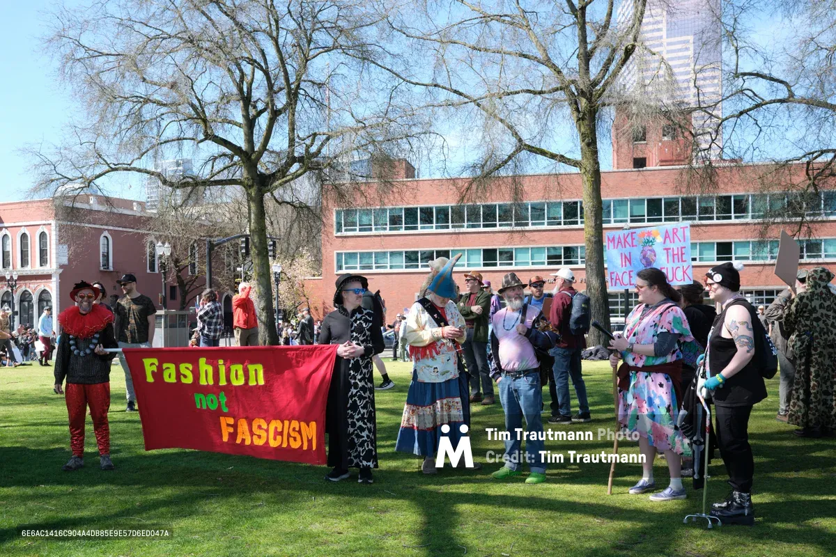 Demonstrators assemble on the sunlit lawn of Portland's Old Town district during a March 2026 anti-monarchy protest, their colorful banners creating striking focal points against the backdrop of red brick institutional buildings. The diverse crowd, dressed in vibrant clothing and costumes, holds signs including "Fashion not Fascism" and "Make Art in the Face of Muck," while bare-branched trees cast intricate shadows across the green space. The scene captures both the theatrical nature of political expression and the earnest community gathering under clear blue skies.